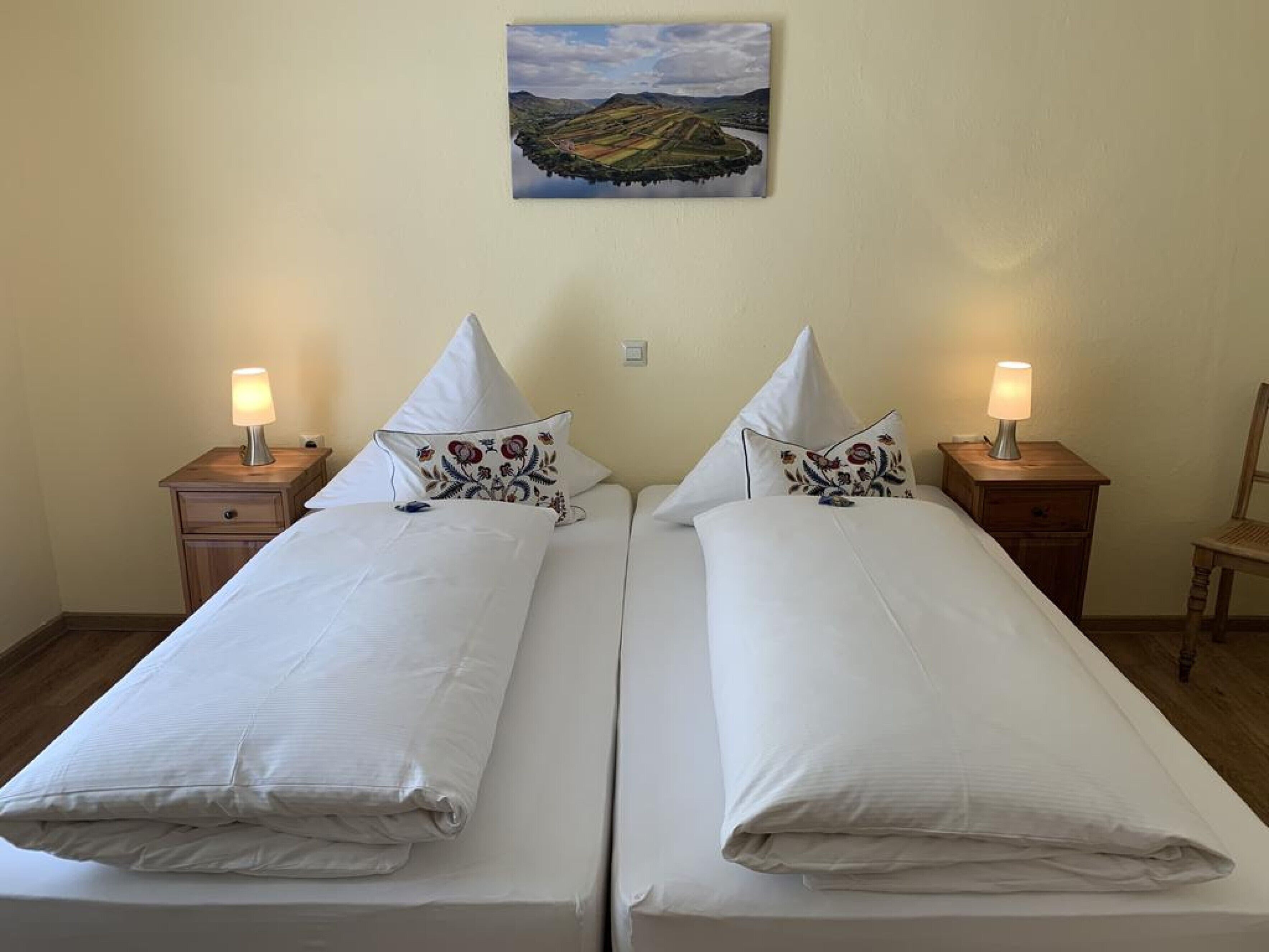 Ein Doppelbett mit weißen Leinentüchern und zwei Zierkissen, begleitet von Nachttischen mit Lampen, in einem Zimmer mit einem Bild an der Wand.