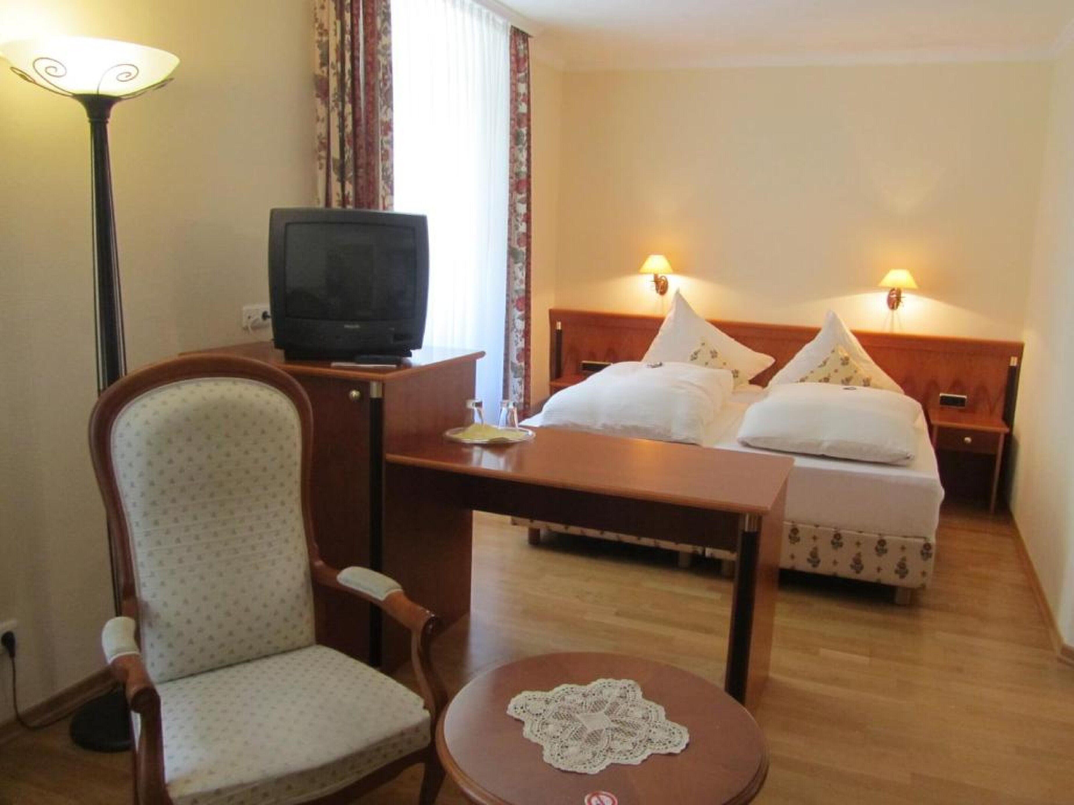 Hotelkamer met een eenpersoonsbed, bureau met televisie, stoel en een klein tafeltje met een tafelkleedje.