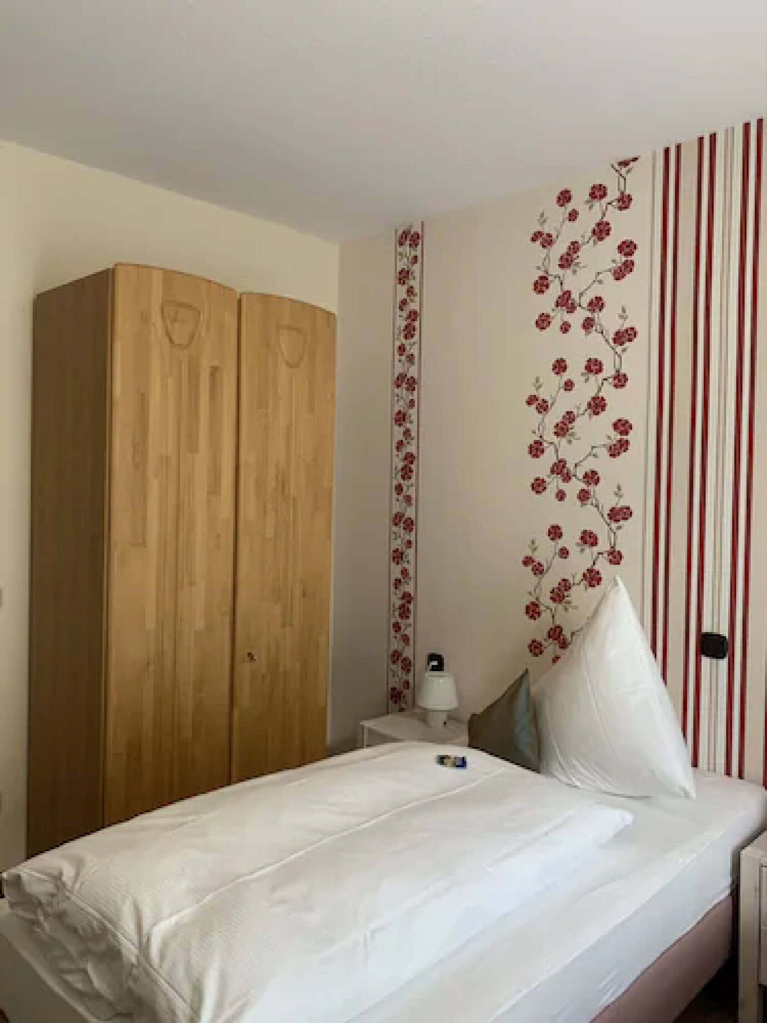 Un dormitorio con un armario de madera, una cama con ropa de cama blanca, una mesita de noche con lámpara y papel tapiz floral.