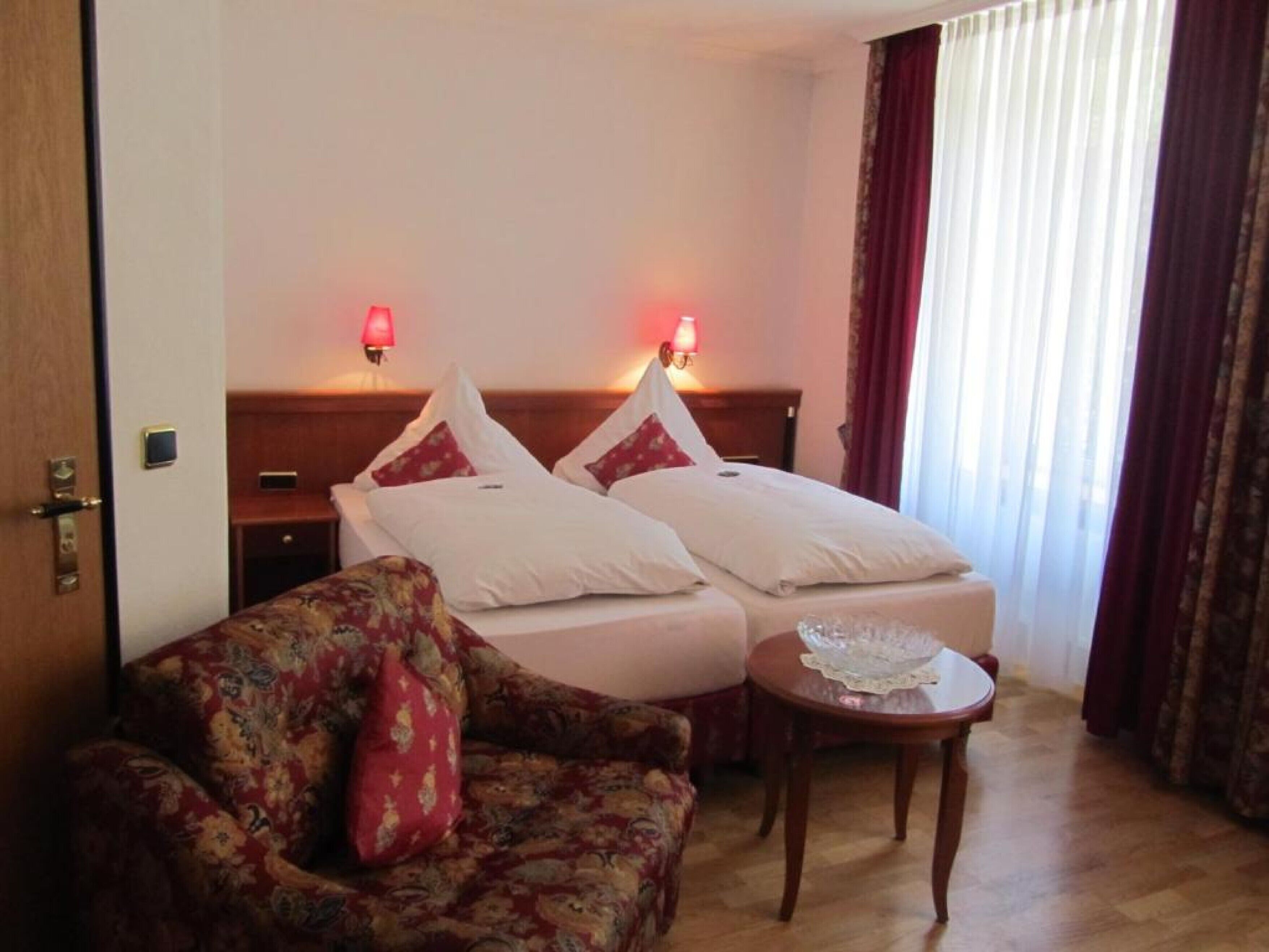 Ein Hotelzimmer mit zwei Betten, einem blumigen Sofa, einem kleinen Tisch und Blick durch transparente Vorhänge nach draußen.