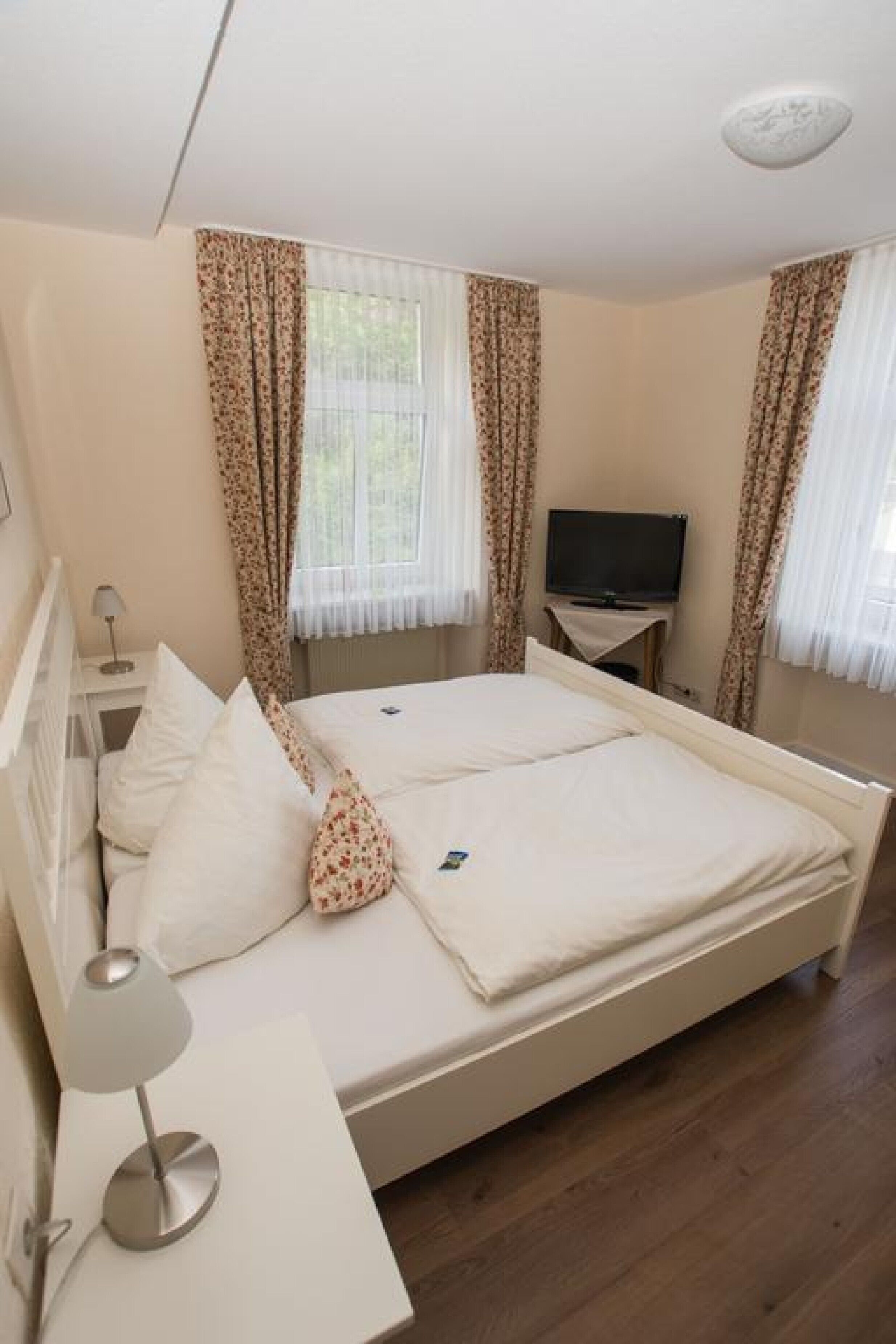 A bedroom with a bed, a TV on a stand, a bedside table with a lamp, and curtains.
