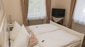 A bedroom with a bed, a TV on a stand, a bedside table with a lamp, and curtains.