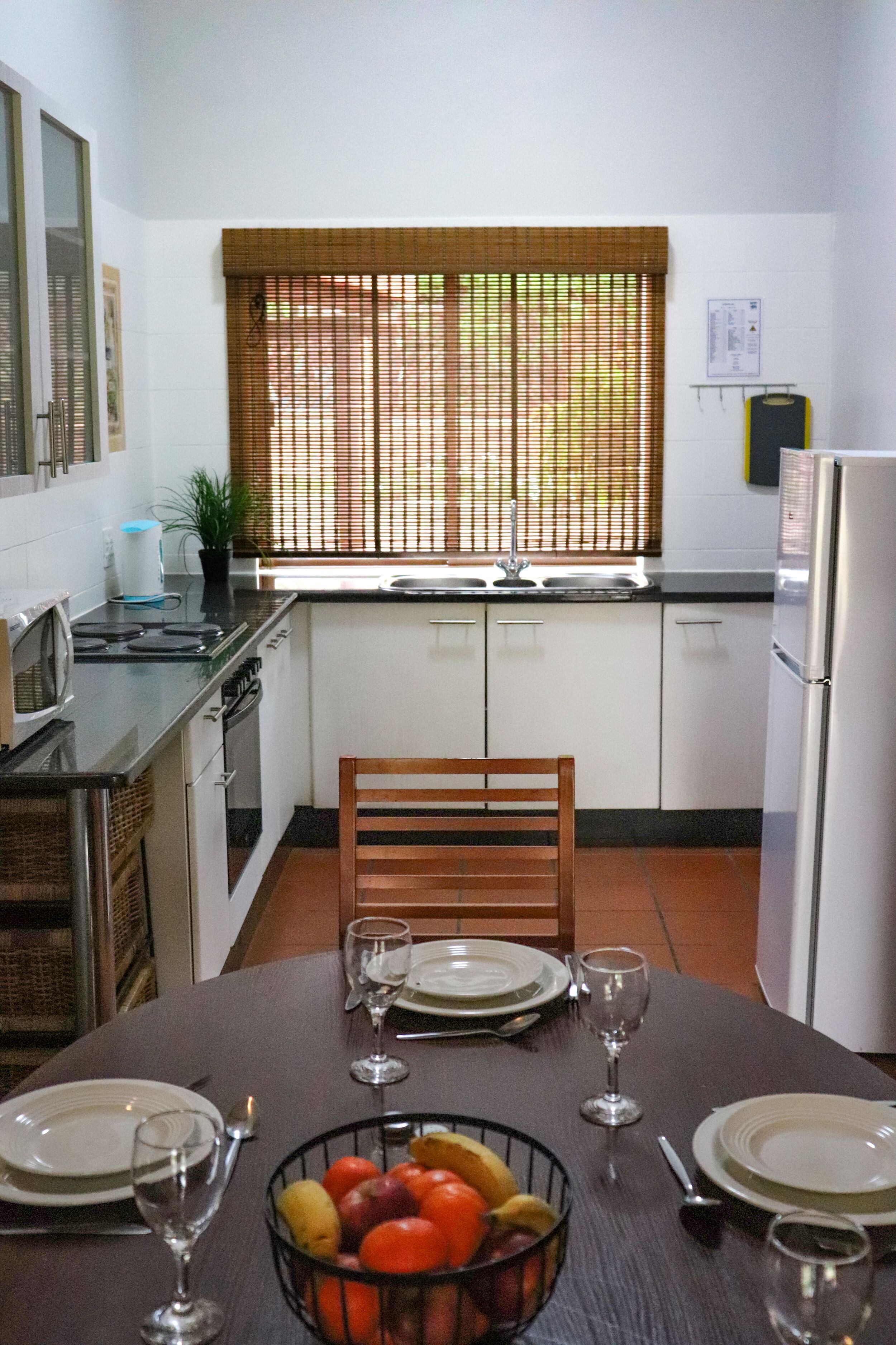 Une cuisine avec une table à manger dressée pour deux personnes, un panier de fruits et un réfrigérateur.