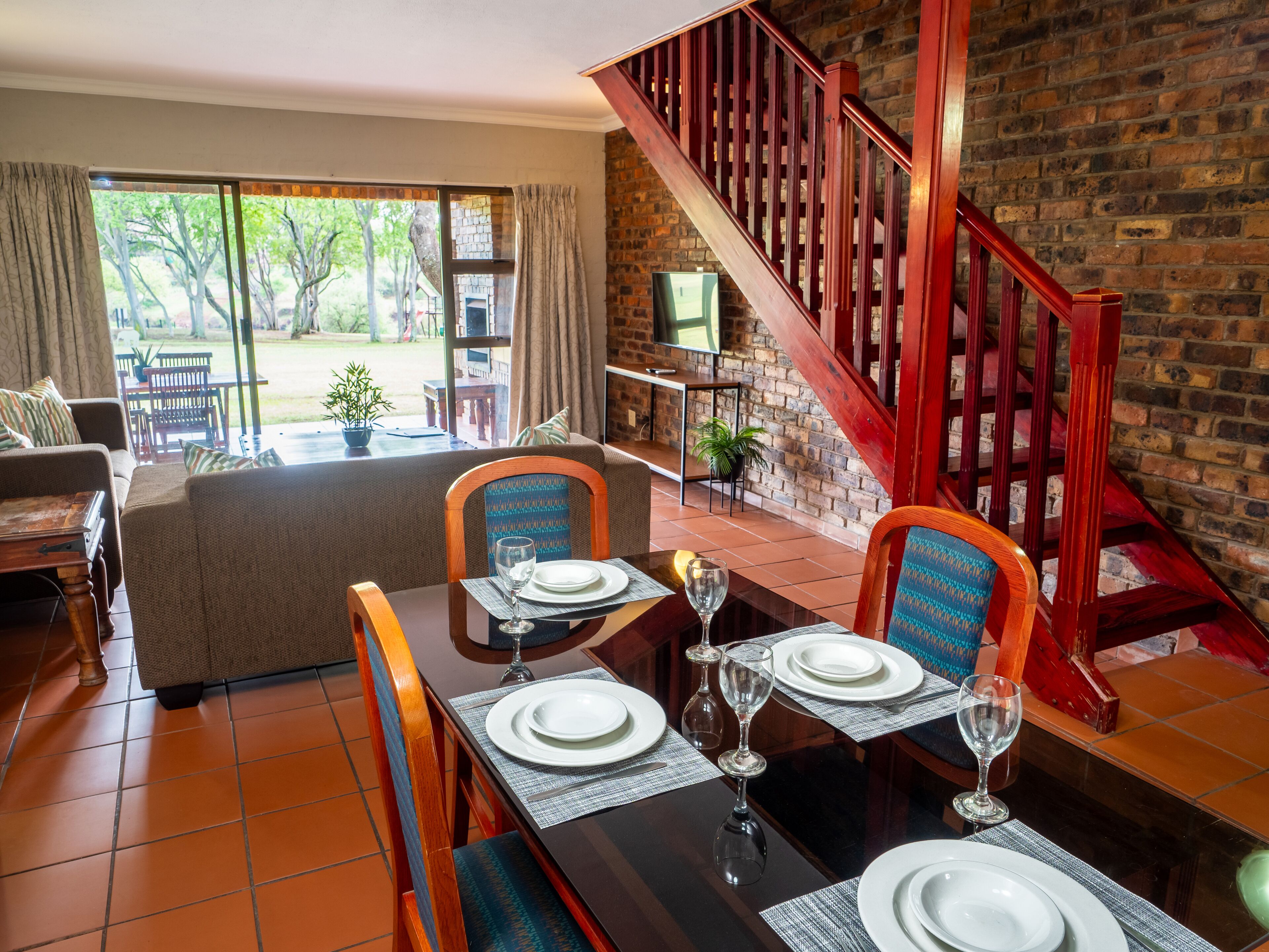 A dining area with a wooden table set for four, a staircase with red railings, and a brick wall.