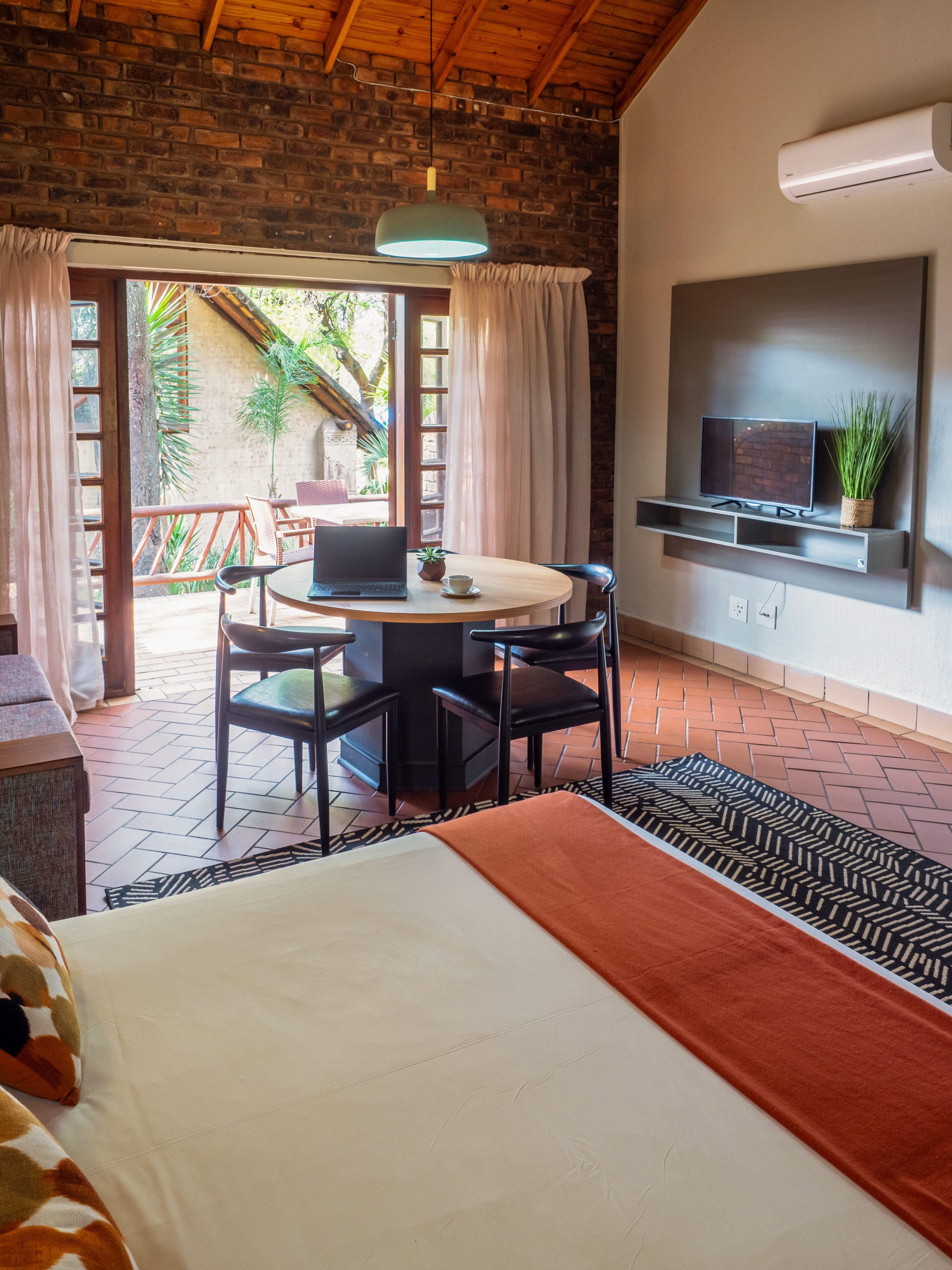 A bedroom with a bed, a desk with a laptop, a TV mounted on the wall, and a balcony with a view of greenery.