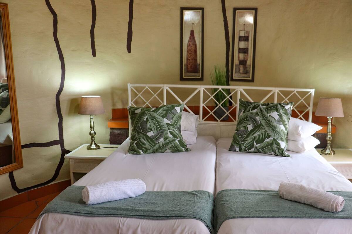 A hotel room with two beds, a white headboard, a nightstand with a lamp, and a wall with two framed pictures.