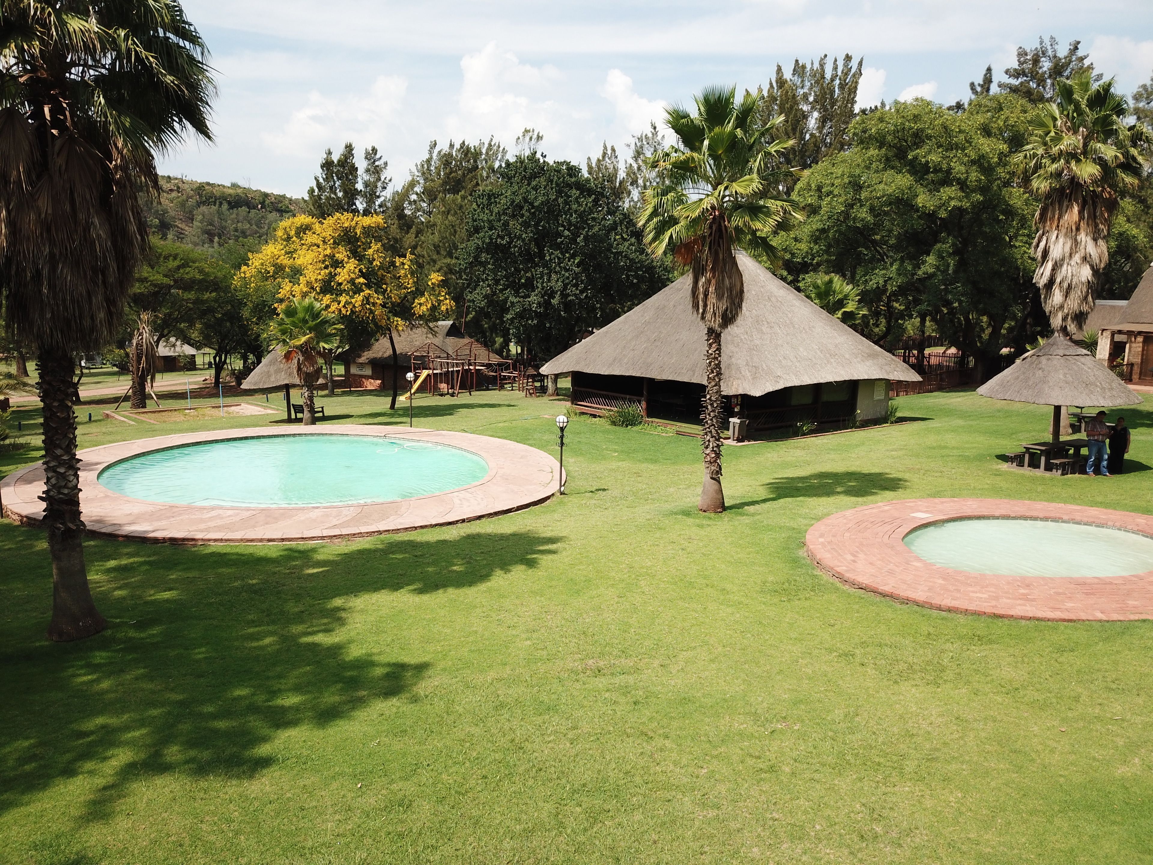 A pool area with two circular pools, surrounded by grass and palm trees, with thatched-roof structures in the background.