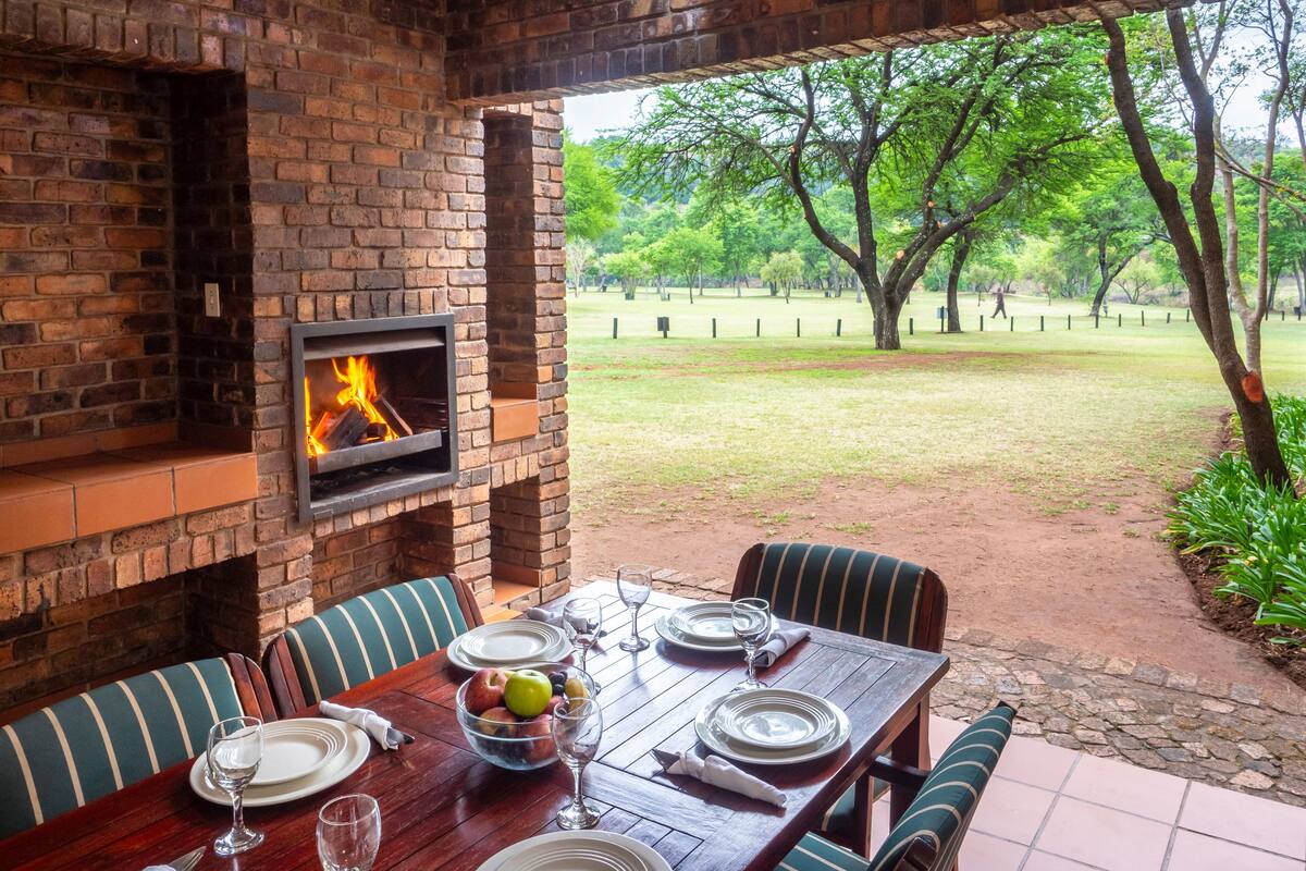 A brick fireplace with a fire, a dining table set with plates and glasses, and a view of a grassy area with trees.