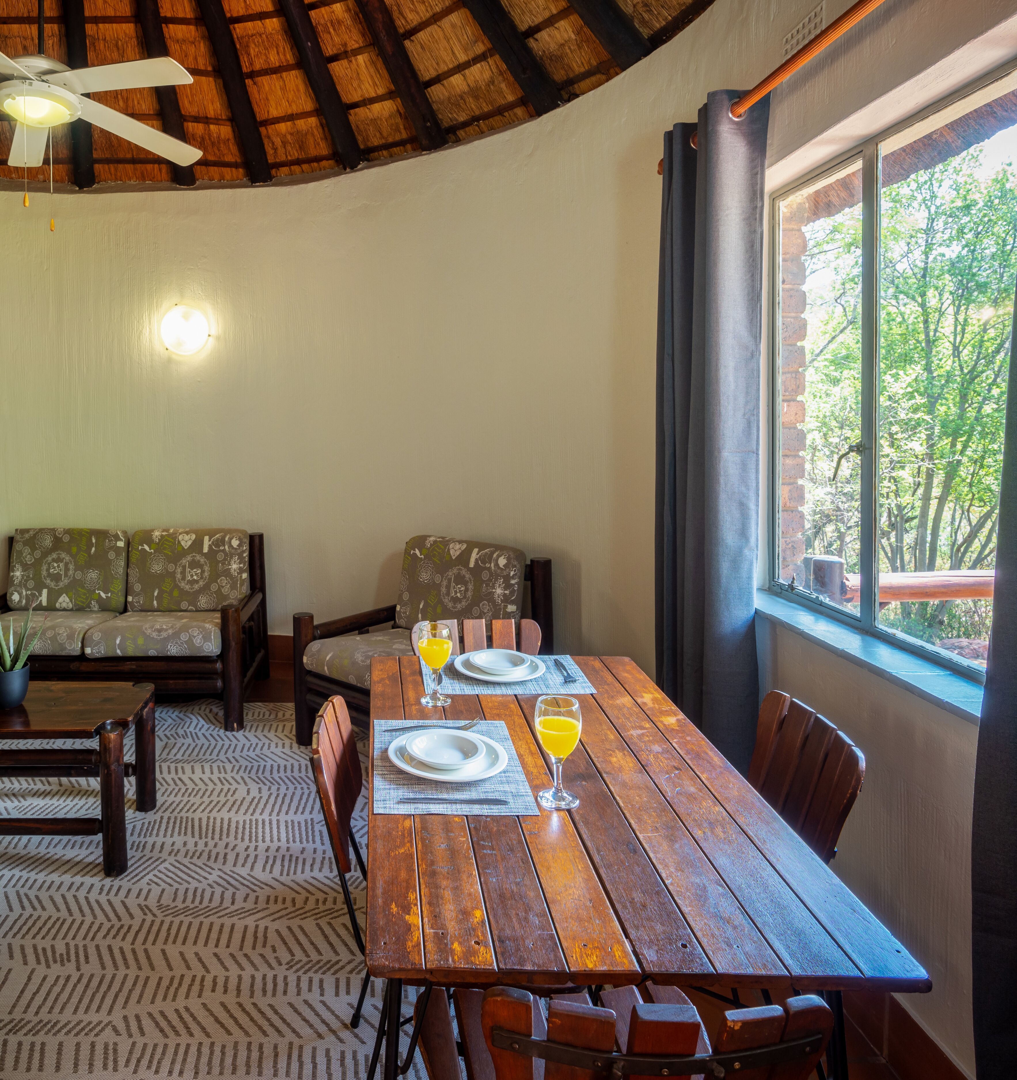 Un área de comedor con una mesa de madera preparada para dos personas, un ventilador de techo y una ventana con vista a árboles.