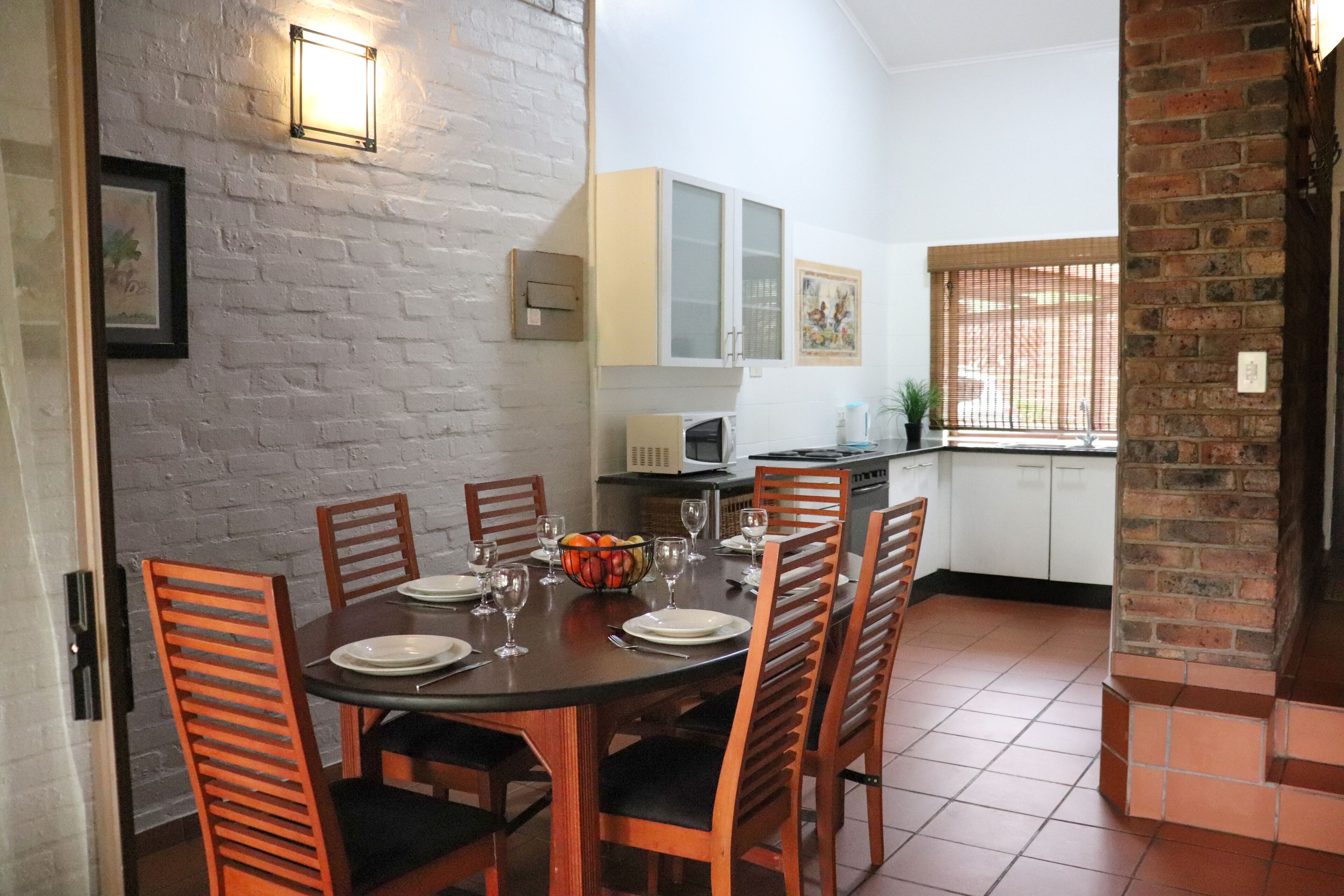 A dining area with a brick wall, a white-framed picture, and a kitchen in the background.