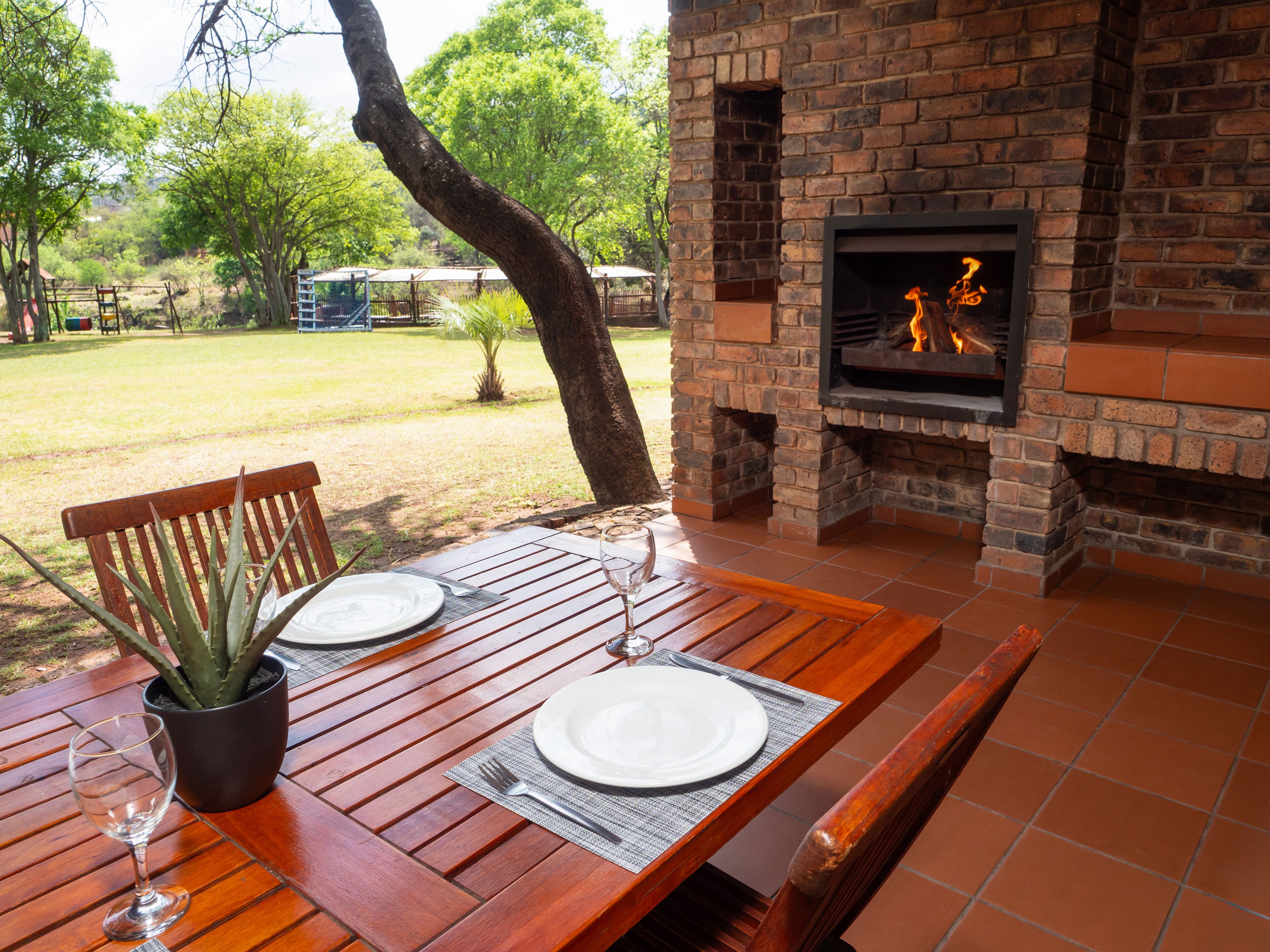 An outdoor dining area with a brick fireplace, wooden table, and chairs.