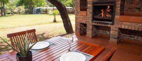 An outdoor dining area with a brick fireplace, wooden table, and chairs.
