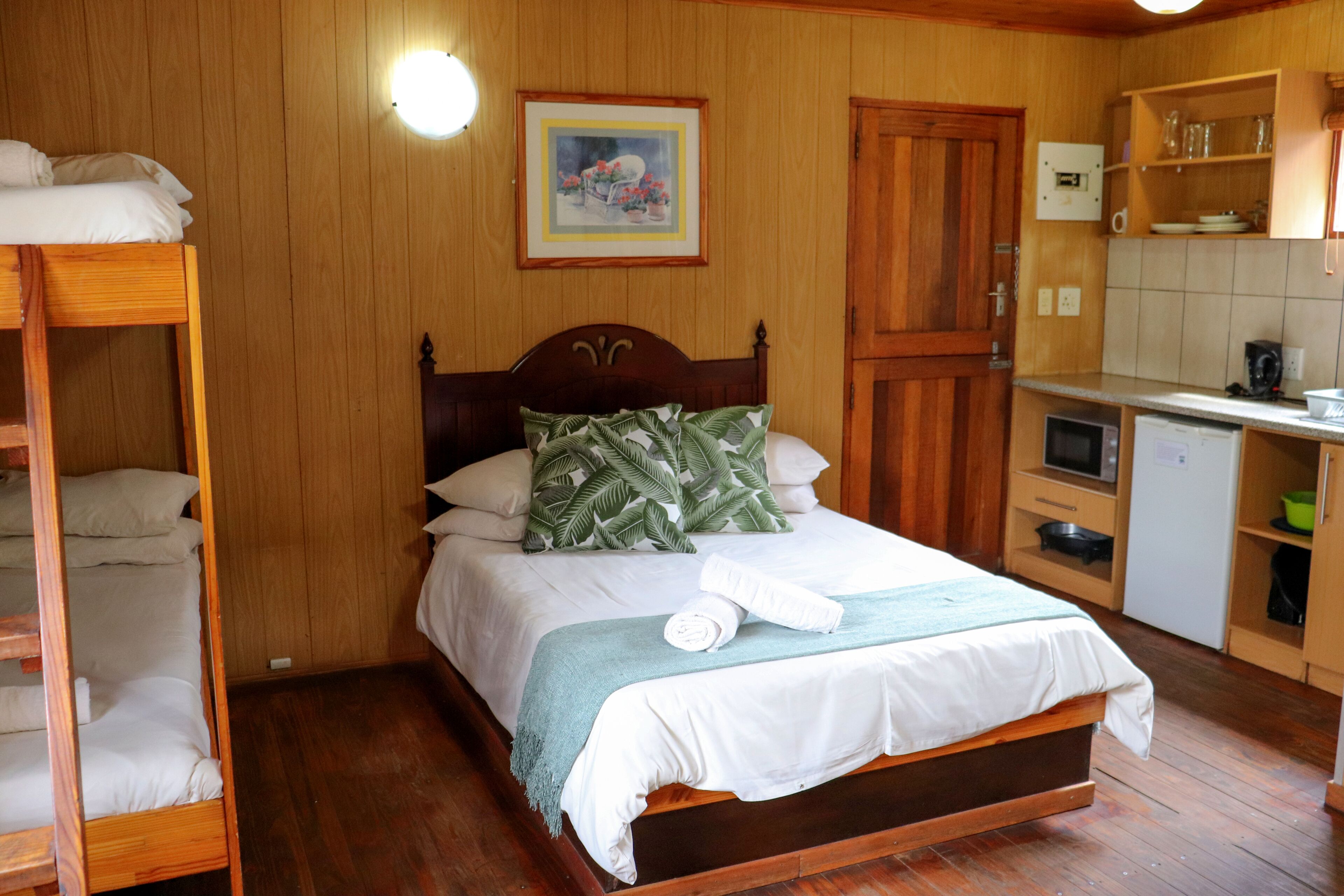 Habitación con paneles de madera, con cama, literas, una zona de cocina y un cuadro en la pared.