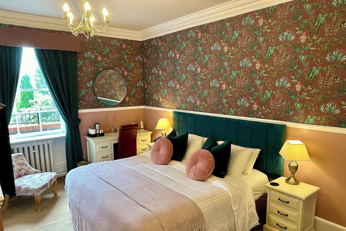 A hotel room with a large bed, a bedside table with a lamp, a chair, a desk with a mirror, and a window with curtains.