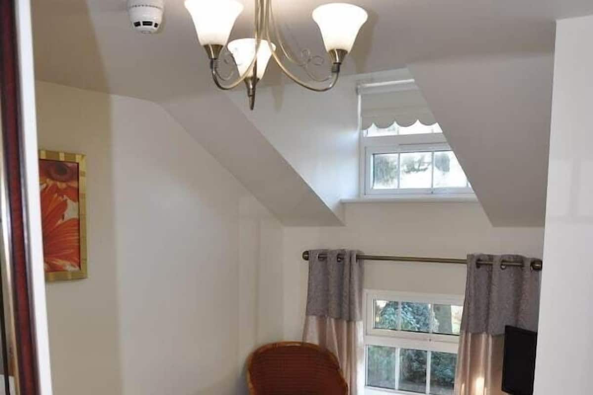 A hotel room with a bed, a chair, a desk with a lamp, a television, a window with curtains, and a ceiling light fixture.