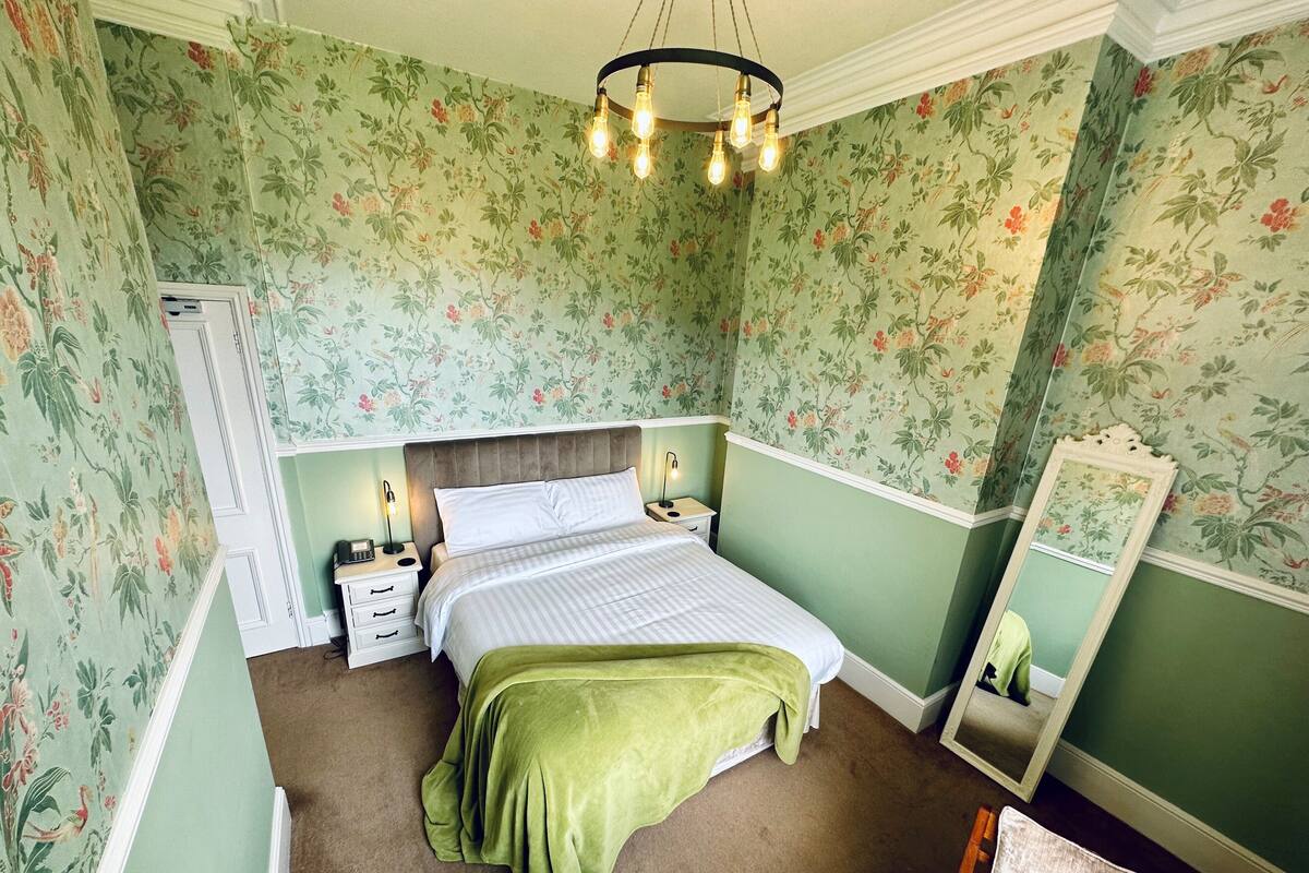 A bedroom with floral wallpaper, a bed with a green blanket, bedside lamps, a nightstand, and a mirror.