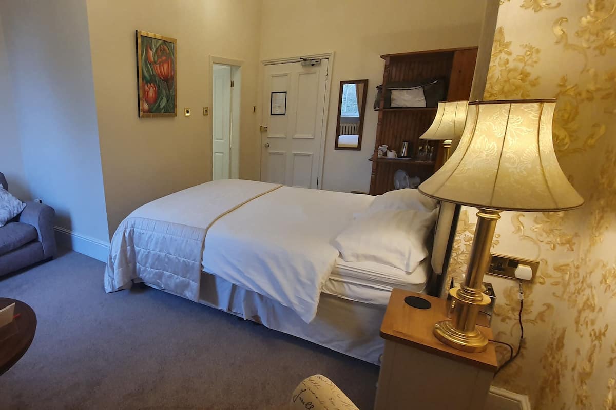 A hotel room with a bed, a sofa, a small table with a remote control, a lamp, and a door leading to another room.