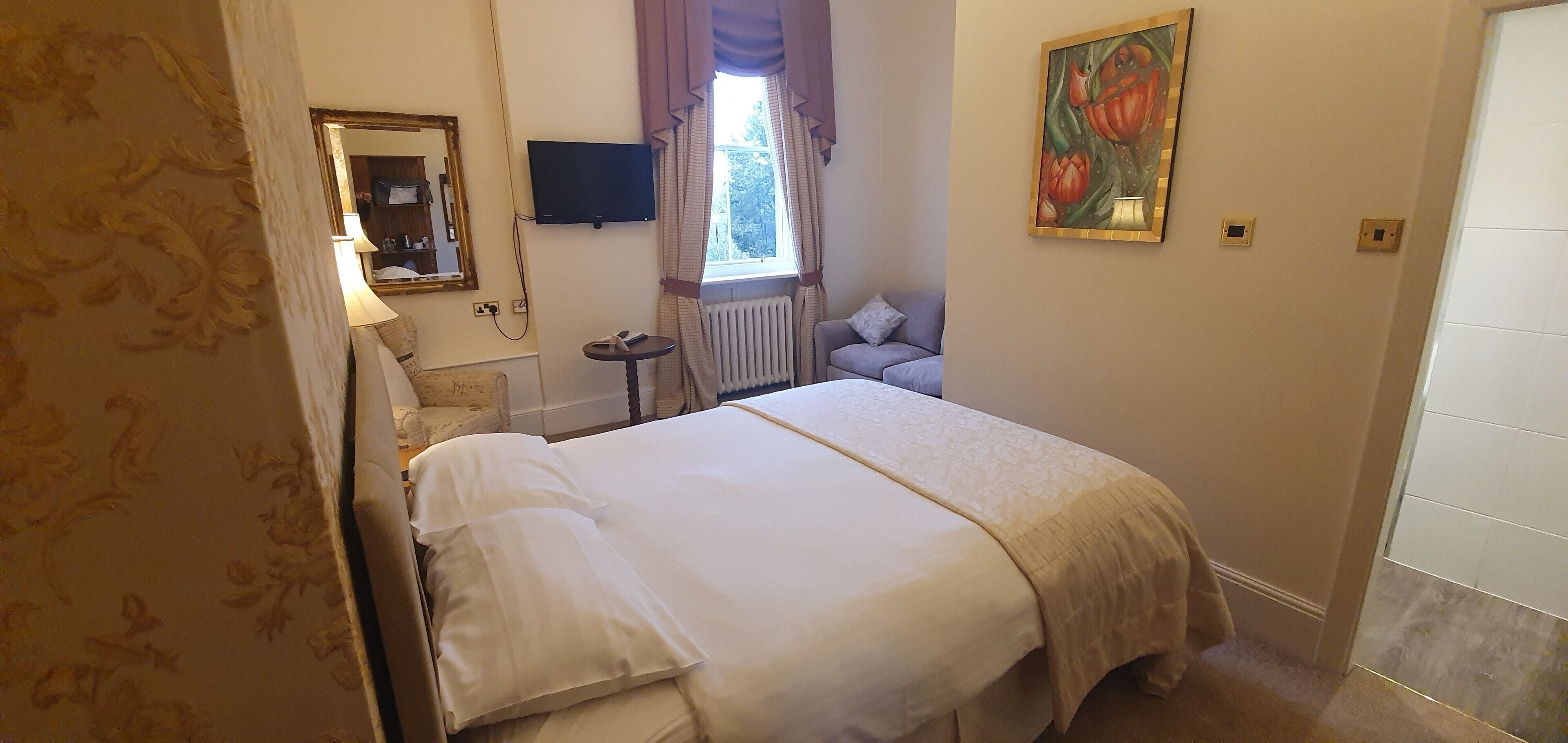 Una camera d'albergo con un letto grande, un tavolino, una sedia, una televisione, una finestra con tende, un termosifone e un quadro appeso al muro.