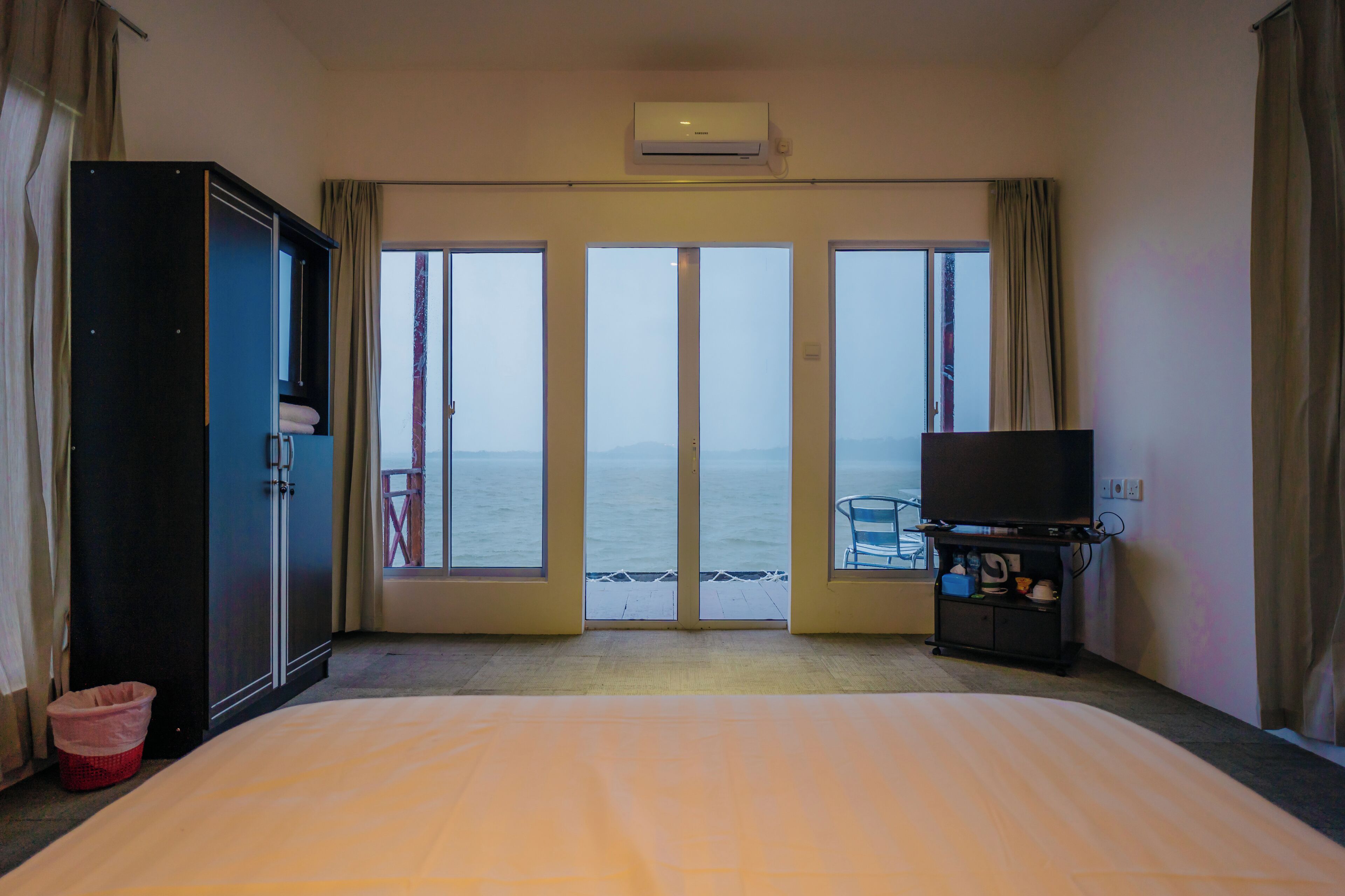 A hotel room with a bed, a TV stand, and a view of the sea through large windows.