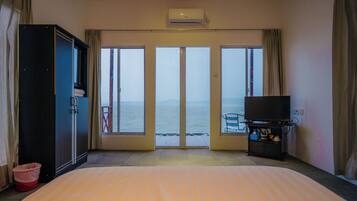 A hotel room with a bed, a TV stand, and a view of the sea through large windows.