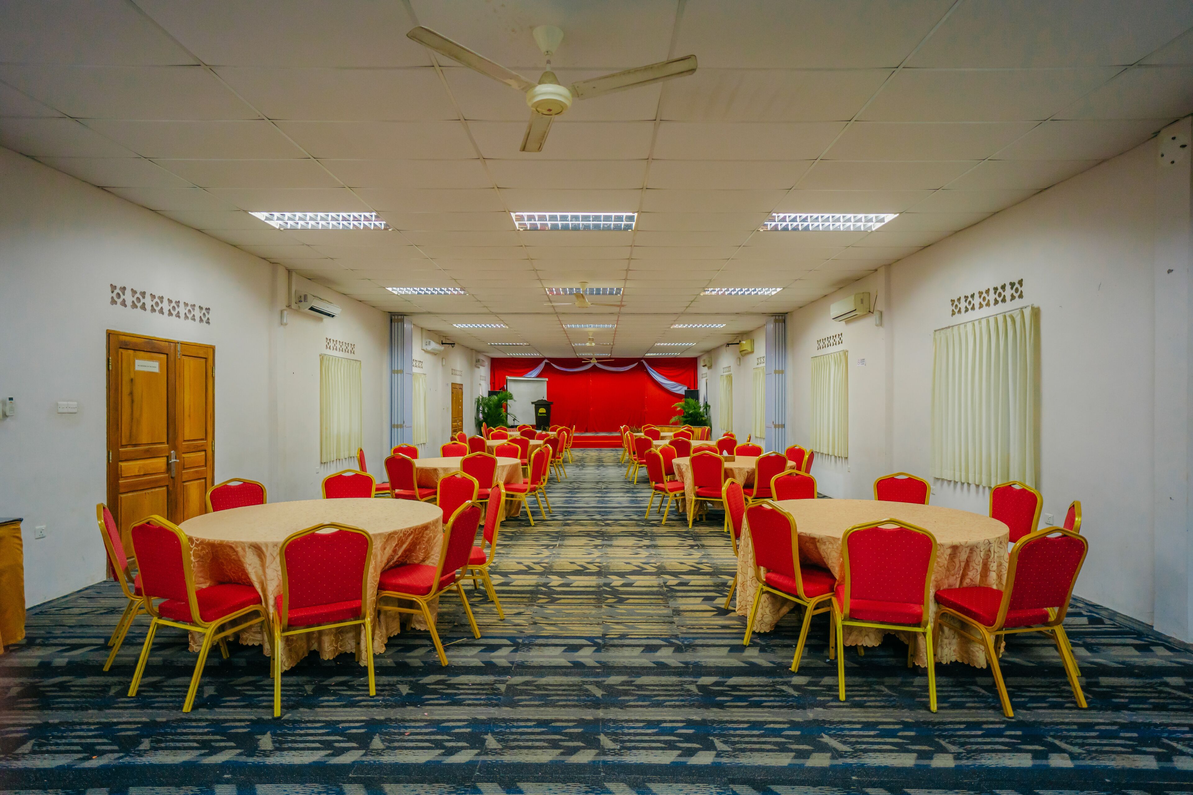 A conference room with multiple tables and chairs, a red carpeted floor, ceiling fans, and fluorescent lighting.