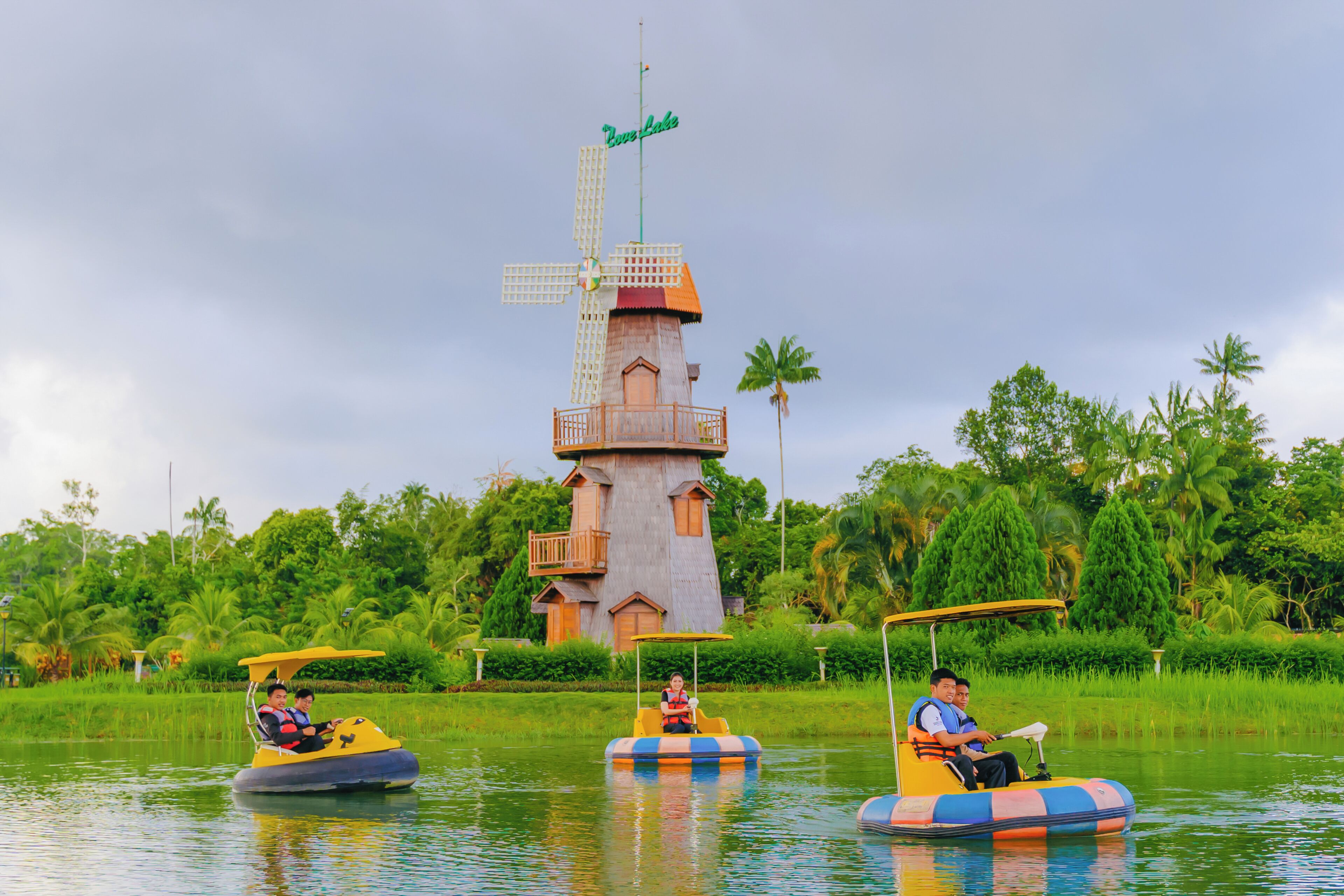 A water ride with colorful inflatable boats, a windmill, and lush greenery in the background.