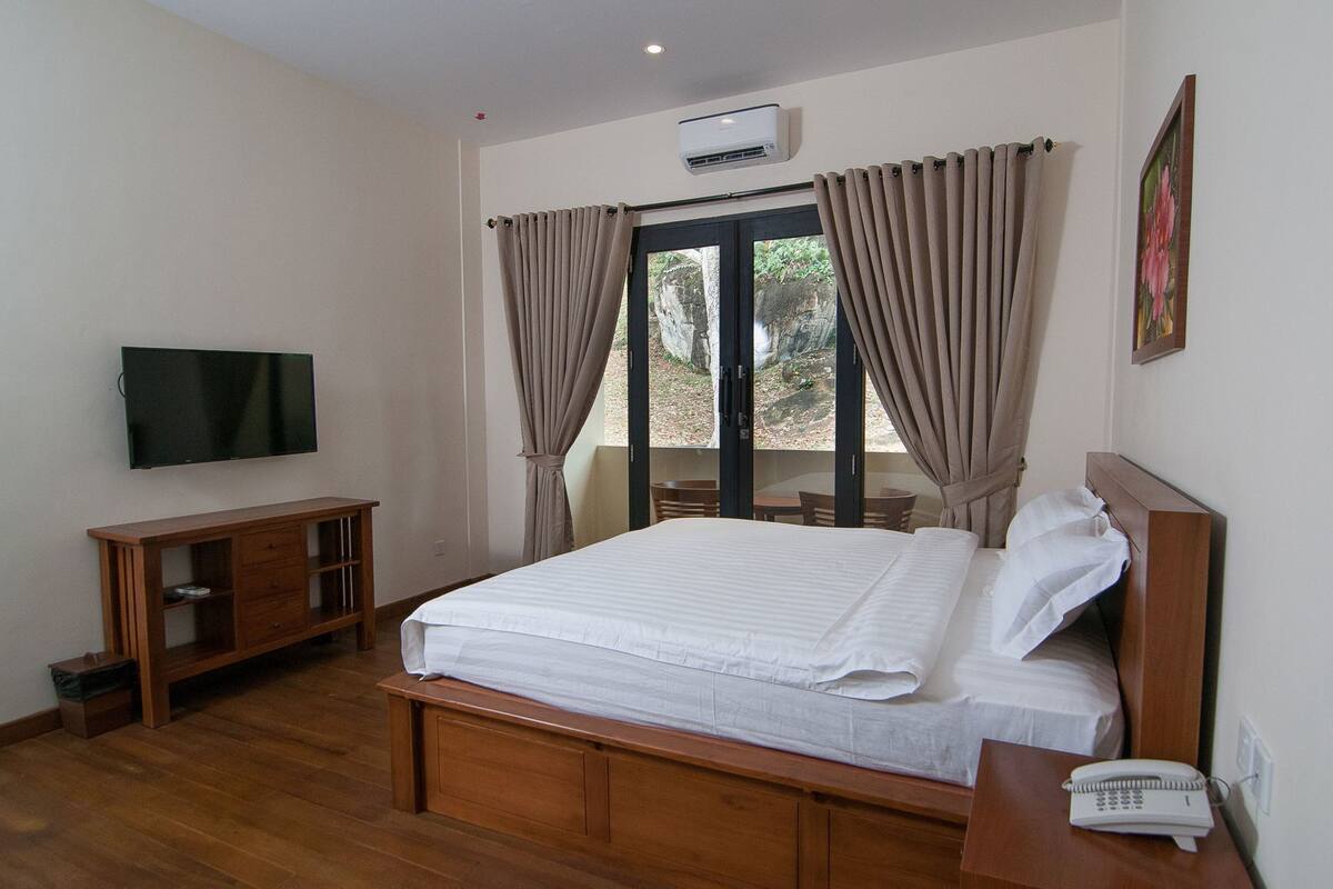 Un dormitorio con cama, televisión, aparador de madera, teléfono y ventana con cortinas.