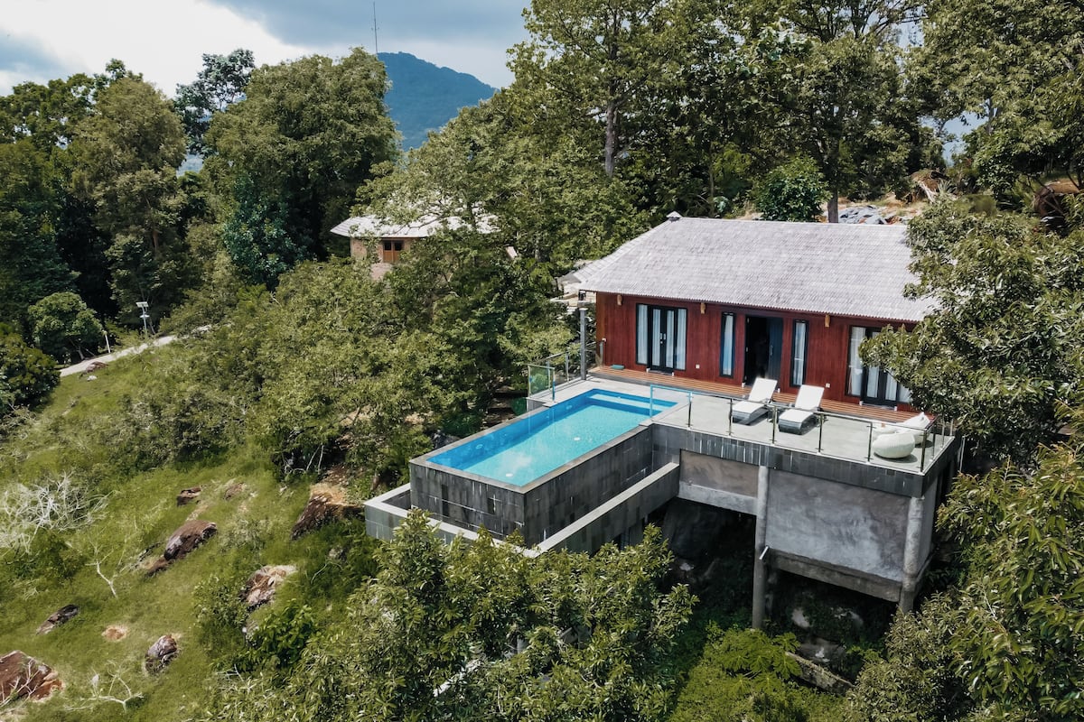 Una casa moderna con piscina, rodeada de exuberante vegetación y una montaña al fondo.