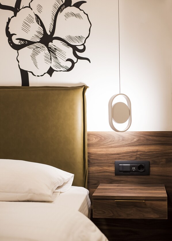 Uma cama bem arrumada com cabeceira, mesa de cabeceira com régua de tomadas e luminária fixada na parede.