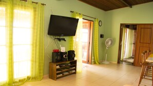 A living room with a television, speakers, and a fan. There is a door leading to another room and a window with curtains. - Chalé Jaguanum (Mangaratiba)