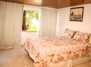 A bedroom with a bed, curtains, a window with a view of greenery, and a painting on the wall. - Chalé Jaguanum (Mangaratiba)