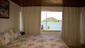 A bedroom with a bed, curtains, a window with a view of a lake and a hill. - Chalé Jaguanum (Mangaratiba)