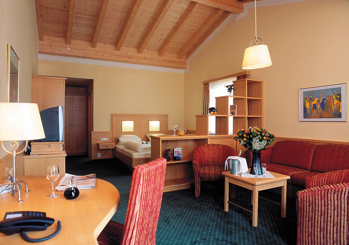 A hotel room with a wooden ceiling, a bed, a desk with a lamp, a red sofa, a small table with a vase, and a painting on the wall.