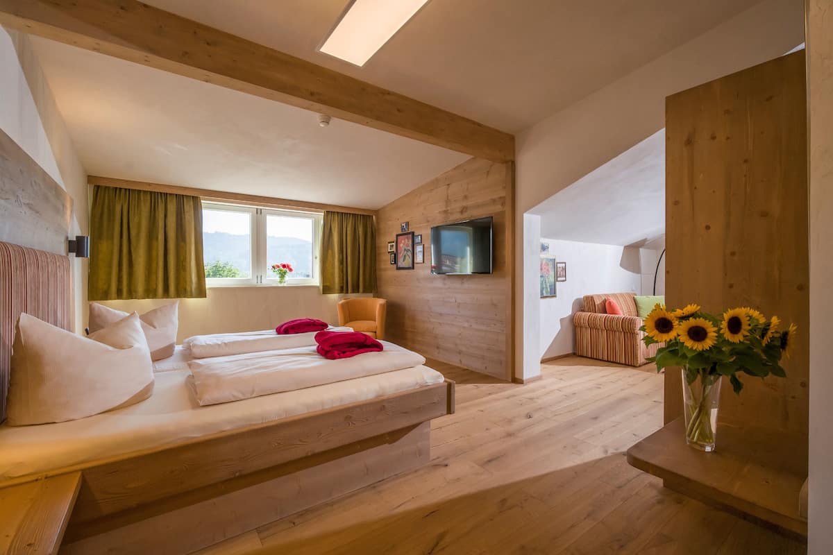 A bedroom with a wooden floor, a bed with white bedding and red pillows, a TV on the wall, and a window with curtains.