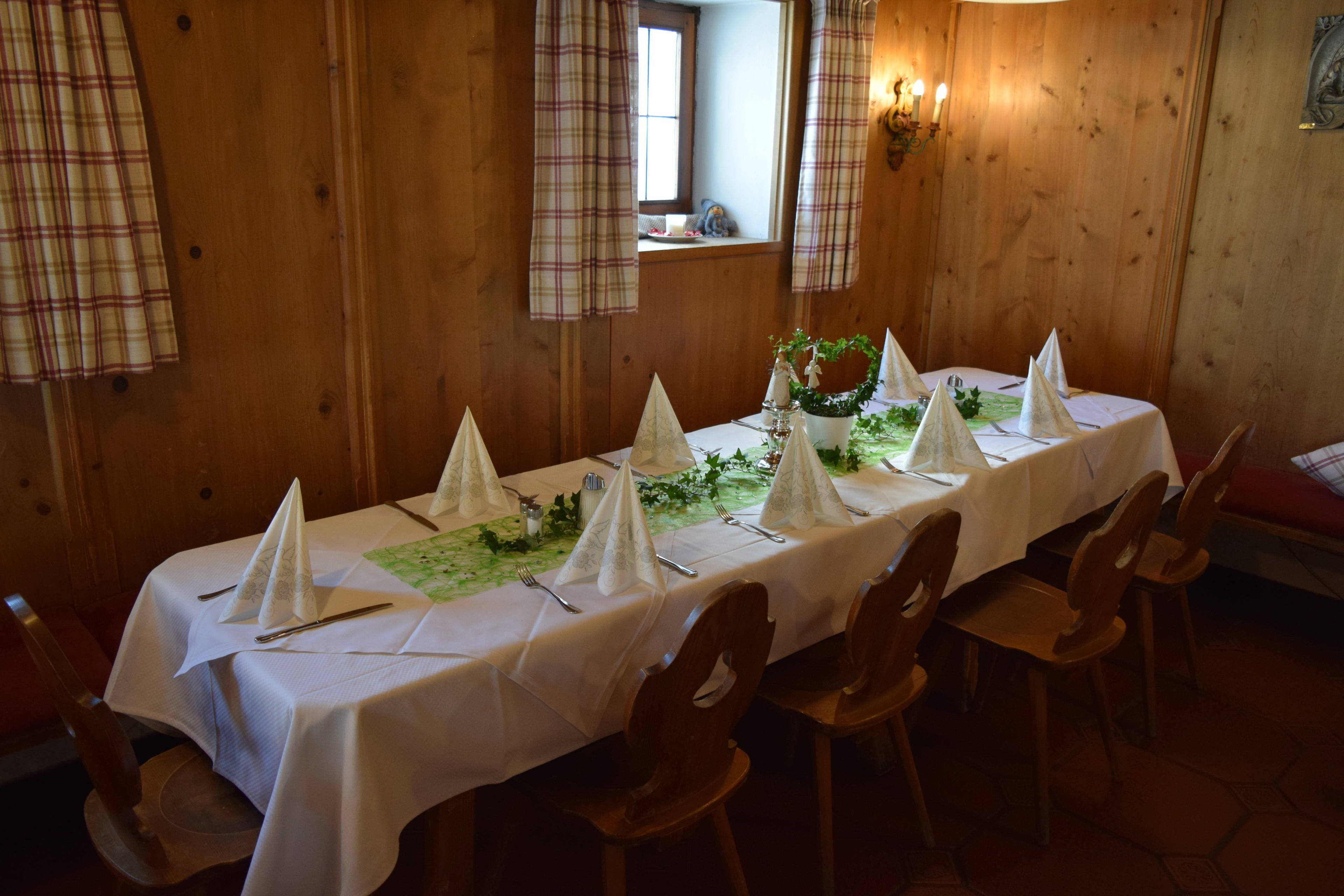 Een lange eettafel gedekt met een witte tafelkleed, servetten en bestek, omringd door houten stoelen in een kamer met houten wanden en een raam.
