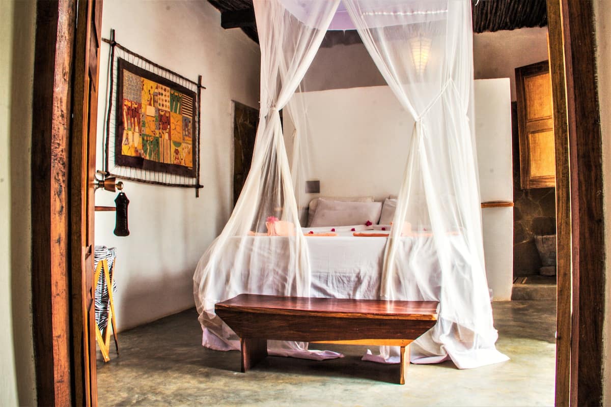 Una habitación con una cama bajo un dosel, un banco, una obra de arte enmarcada en la pared y una puerta de madera.