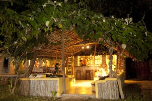 Un área de bar al aire libre con techo de paja, asientos de madera y un sendero de piedra que conduce a la entrada.