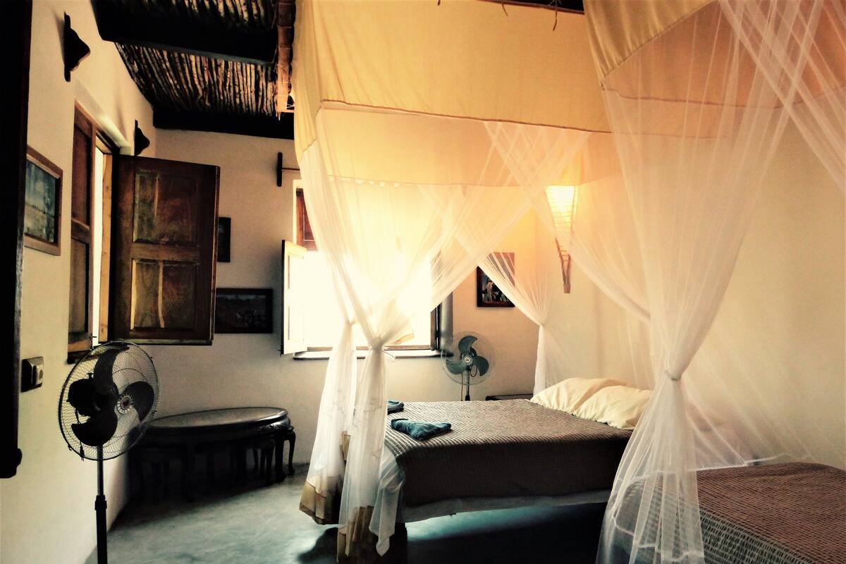 Una habitación con una cama bajo un dosel, un ventilador de pie y una ventana con cortinas.