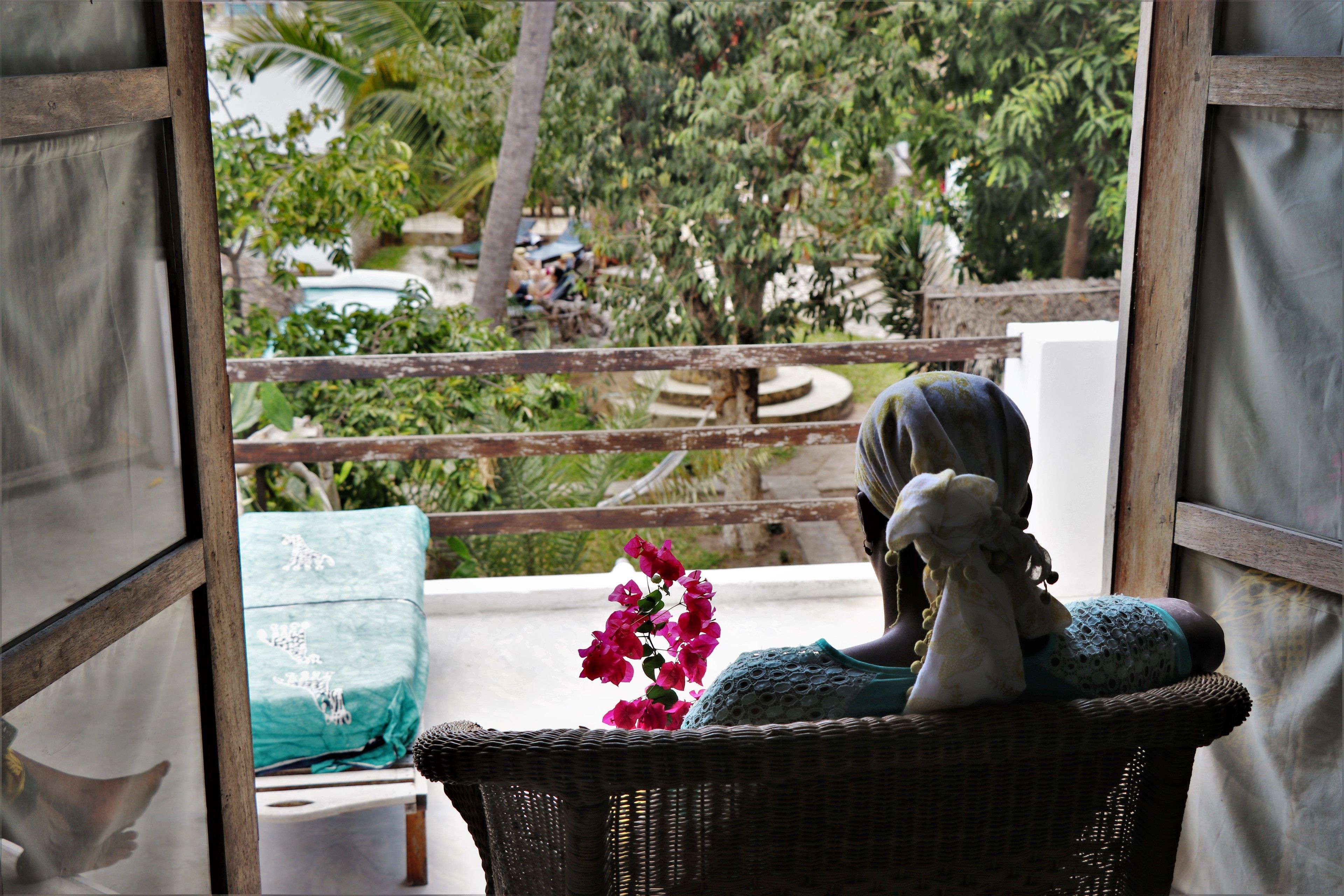 Une chaise en osier agrémentée d’un objet décoratif et d’un coussin vert, placée sur un balcon offrant une vue sur la verdure et une piscine.