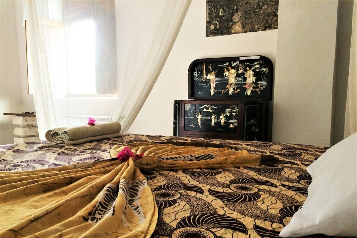 Una cama bien hecha con una colcha estampada, una toalla enrollada y un cojín decorativo.