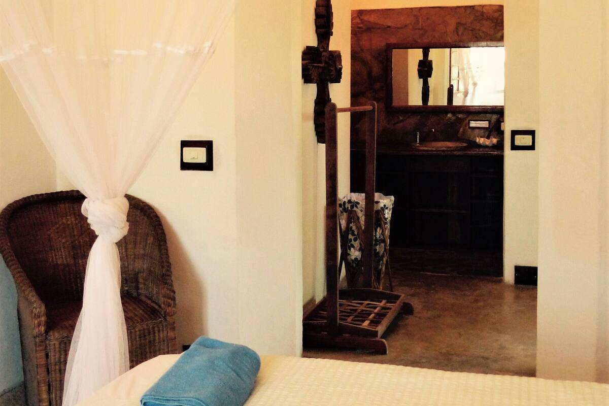 Un dormitorio con cama, una silla, dosel y vista a una zona de cocina.