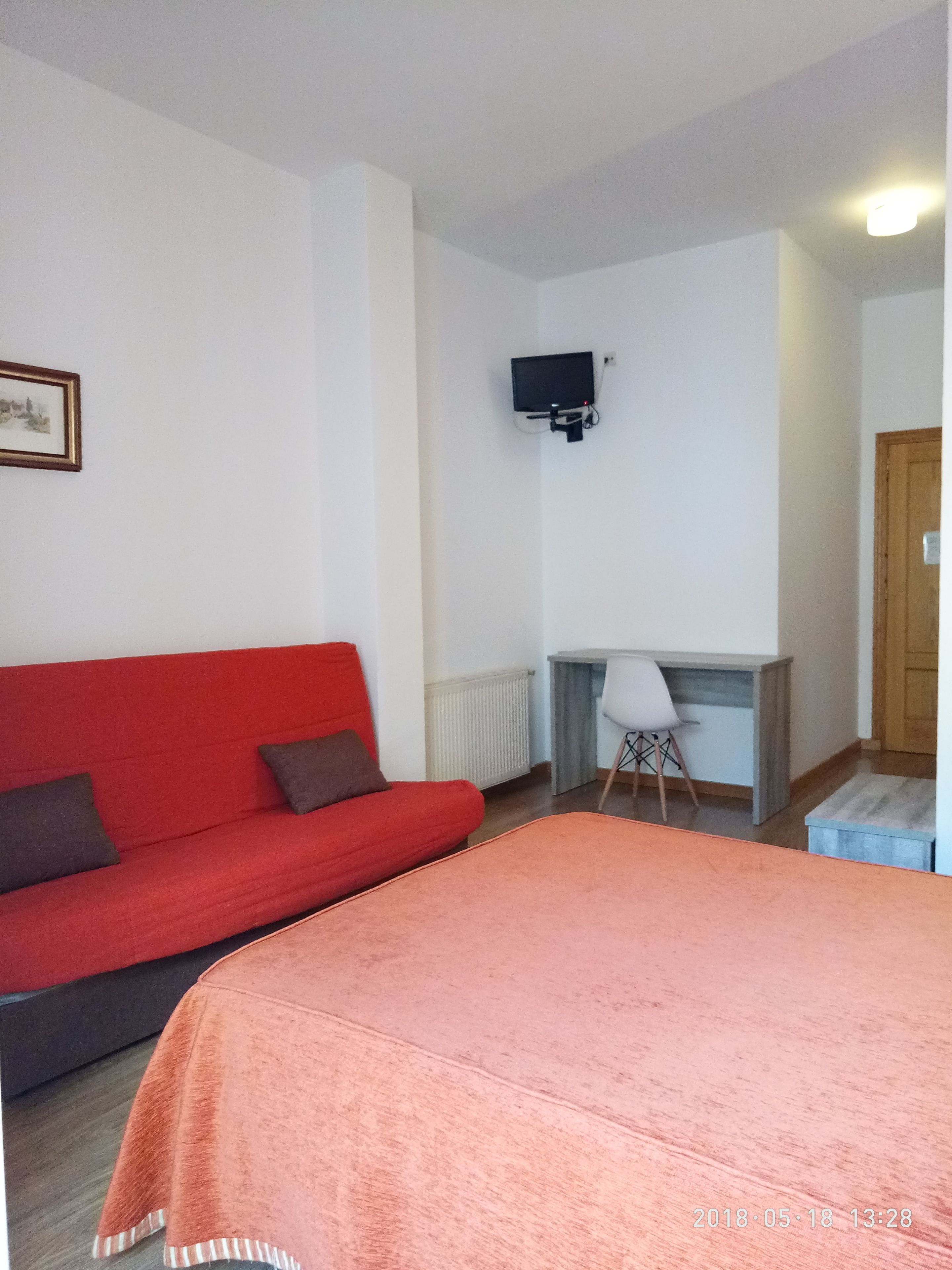 A room with a red sofa, a desk with a chair, a television mounted on the wall, and a wooden door.