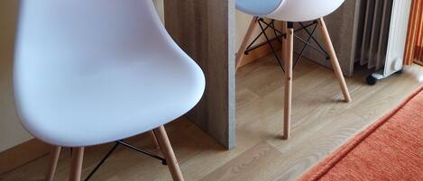 Una habitación pequeña con un escritorio de madera, dos sillas blancas, un radiador y suelo de madera.