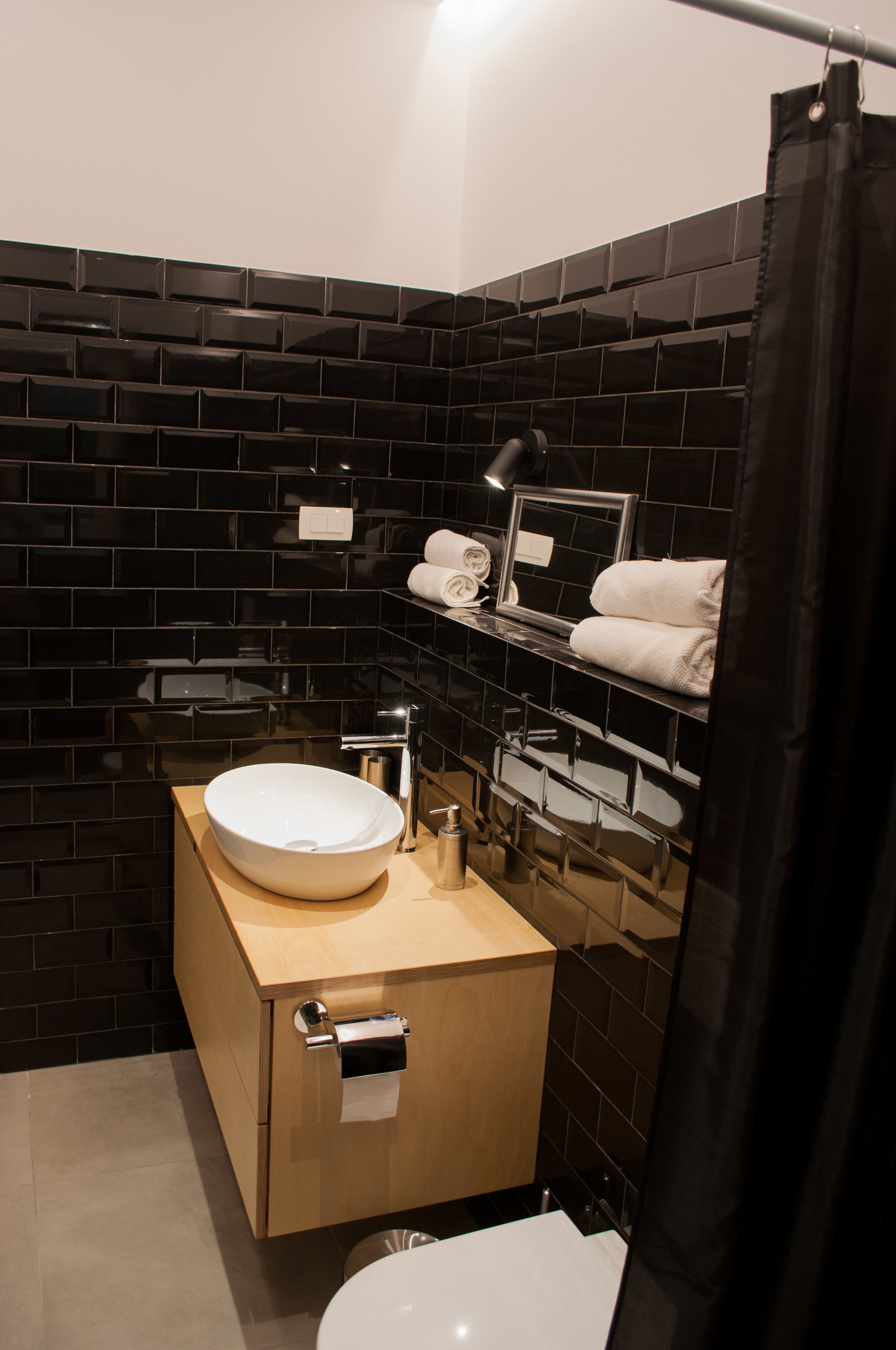 A modern bathroom with a black tiled wall, a wooden vanity, a white sink, a toilet, and a shower curtain.