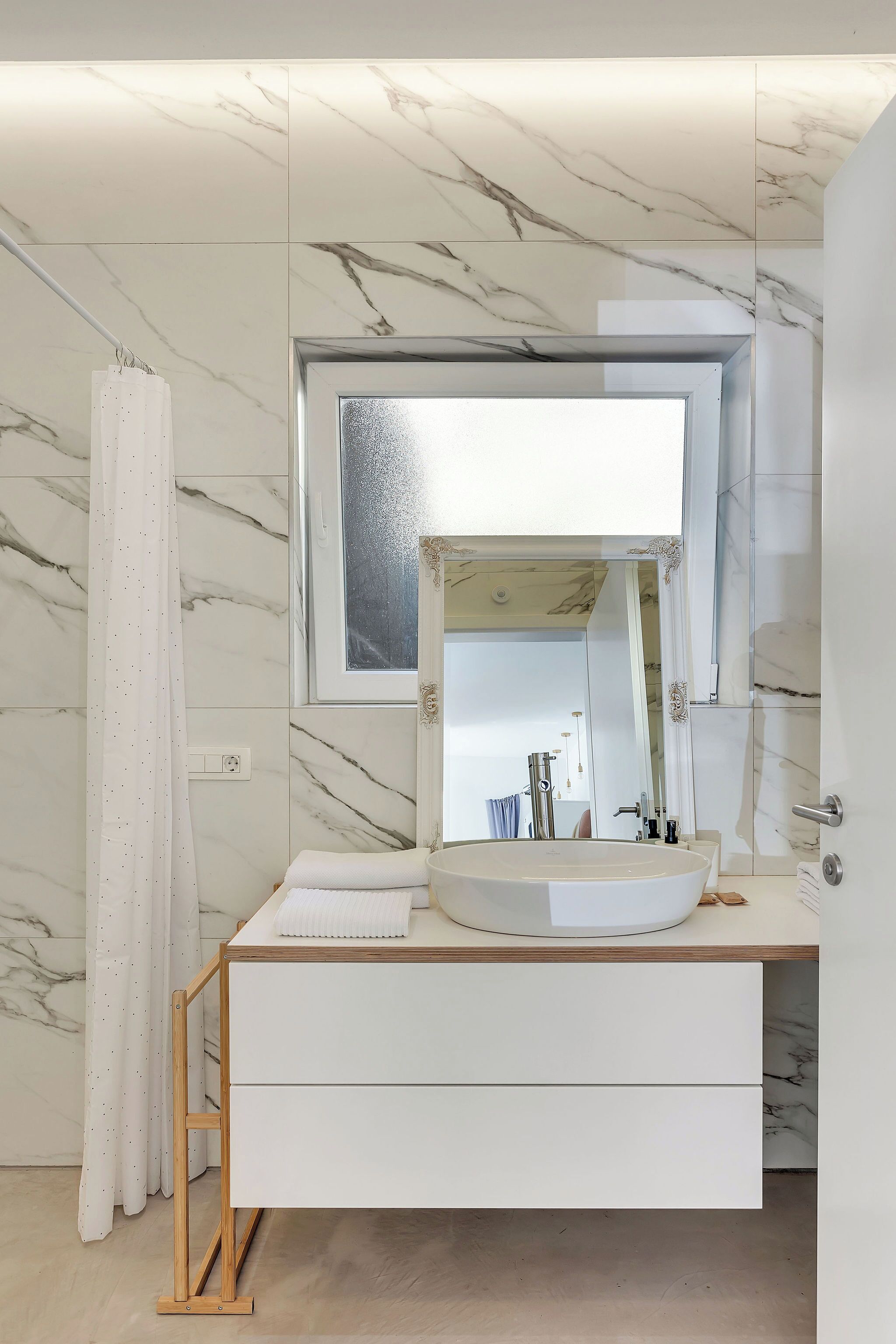 Un baño con pared de mármol, lavamanos blanco y tocador de madera.