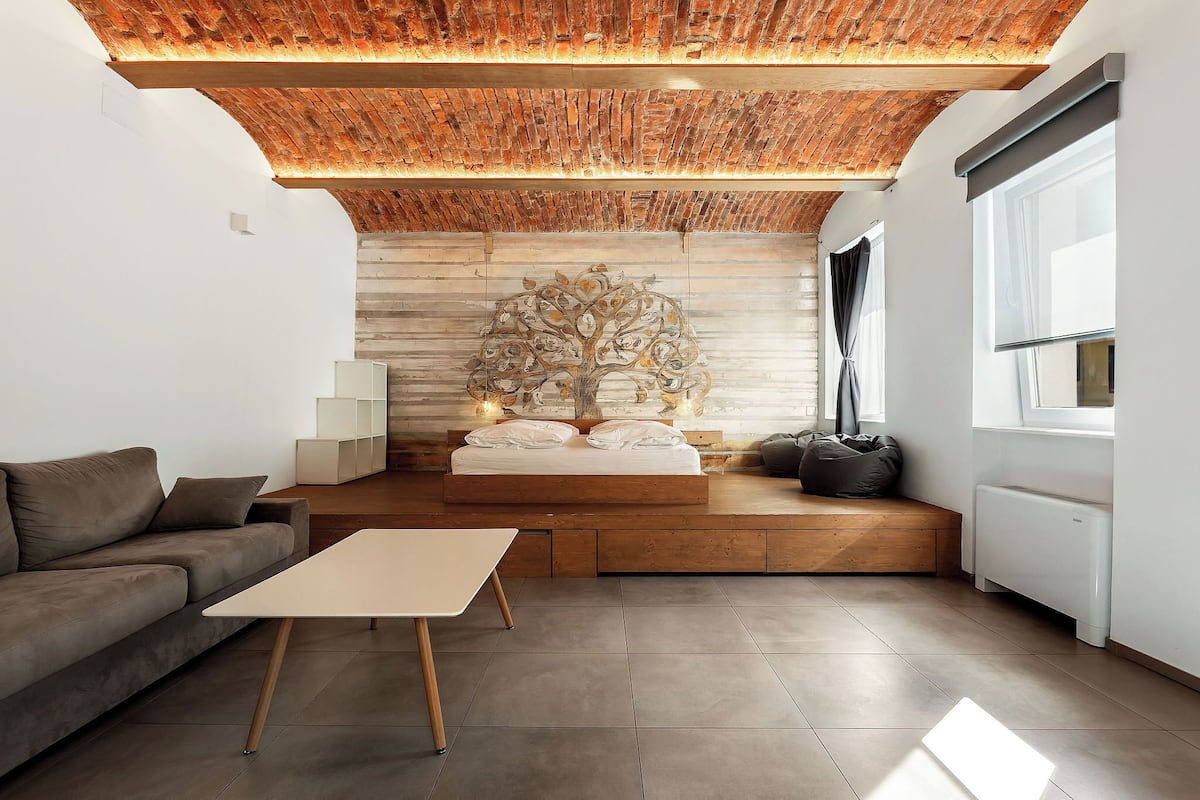 Une chambre moderne avec un plafond en briques, un lit orné d’une fresque représentant un arbre, un canapé, une table de chevet et une fenêtre avec des rideaux.