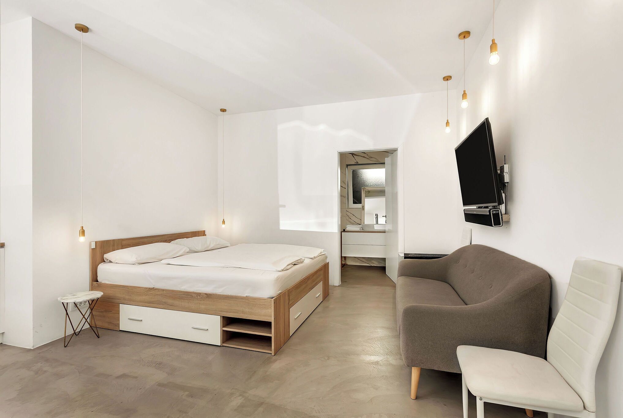A modern bedroom with a wooden bed, a small side table, a sofa, and a TV mounted on the wall.
