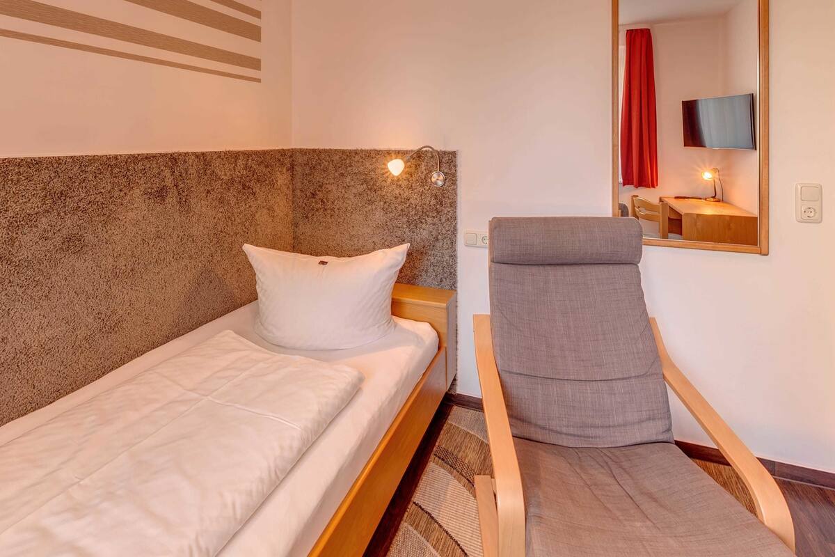 Una cama individual con cabecera, una silla gris, una mesita con lámpara y un espejo en la pared.