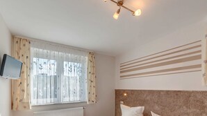 A hotel room with two beds, a mounted TV, a window with sheer curtains, a radiator, and a ceiling light fixture. - Pension Wernesgrün (Steinberg)