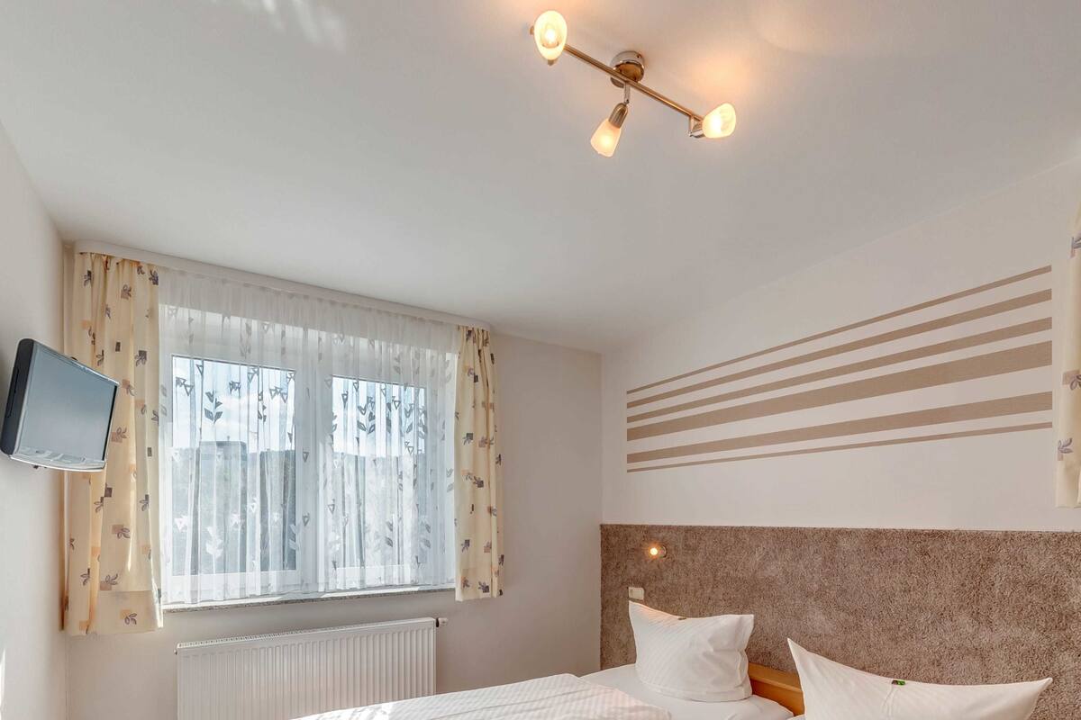 Habitación de hotel con dos camas, un televisor montado, una ventana con cortinas translúcidas, un radiador y una lámpara en el techo.
